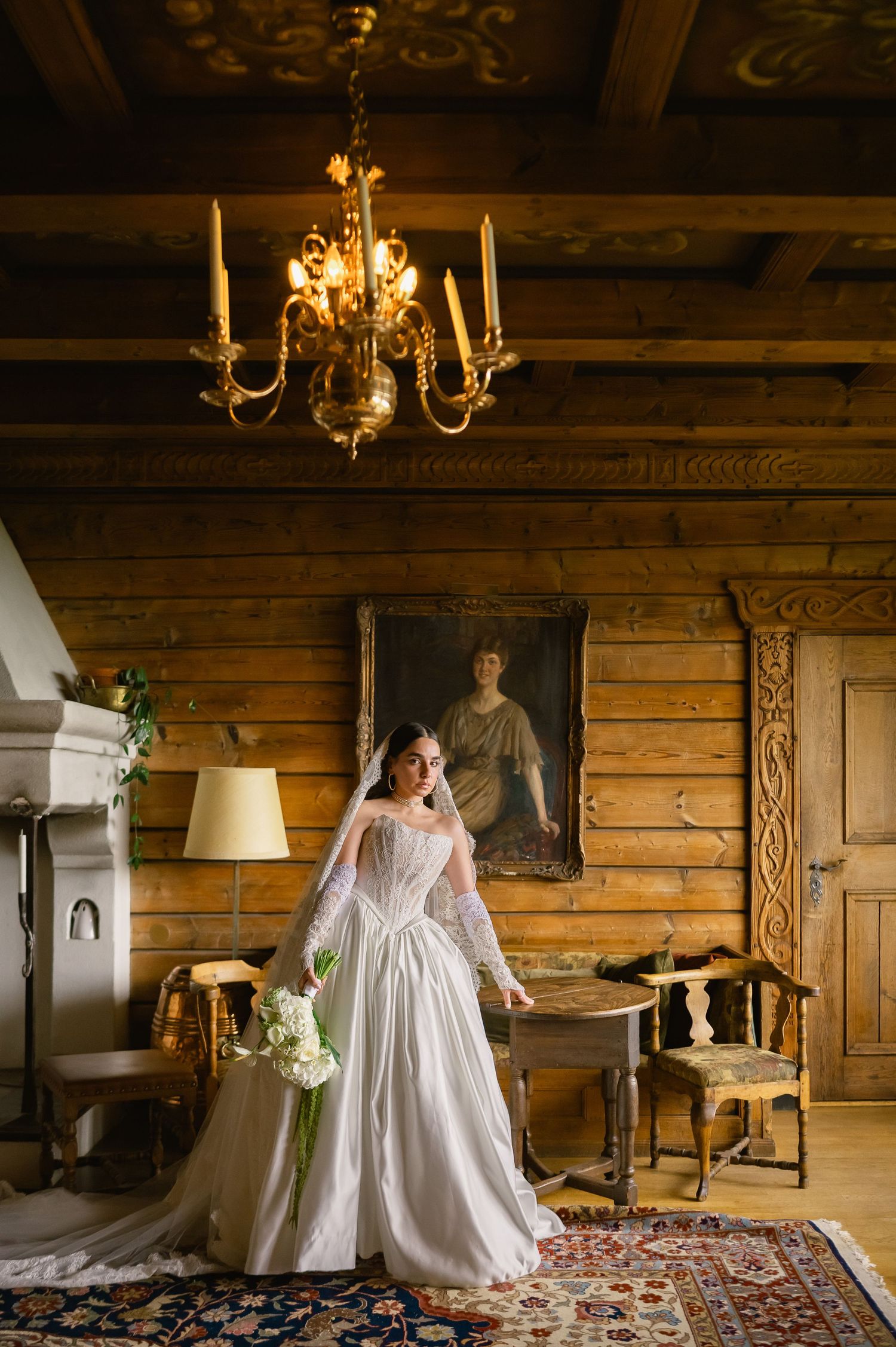 Elegant bride in white gown stands in rustic log cabin room with chandelier and vintage portrait.