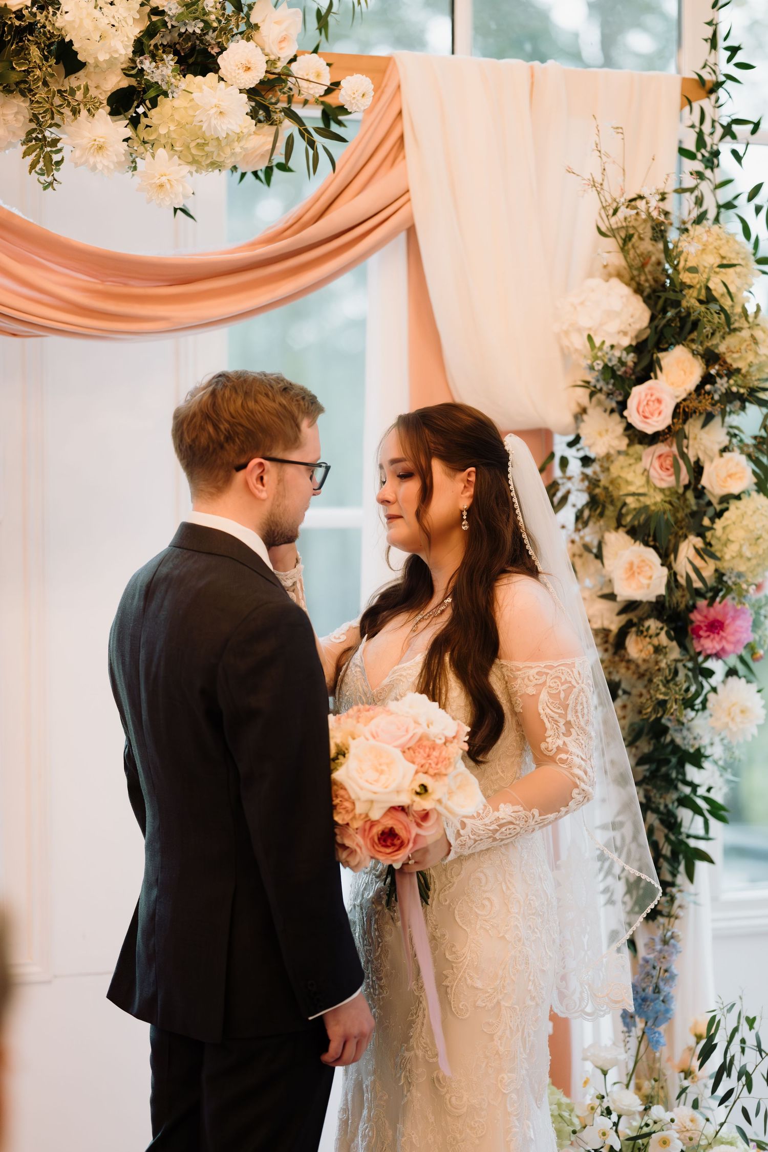 Bride and groom exchange vows under a floral arch with peach draping and roses.
