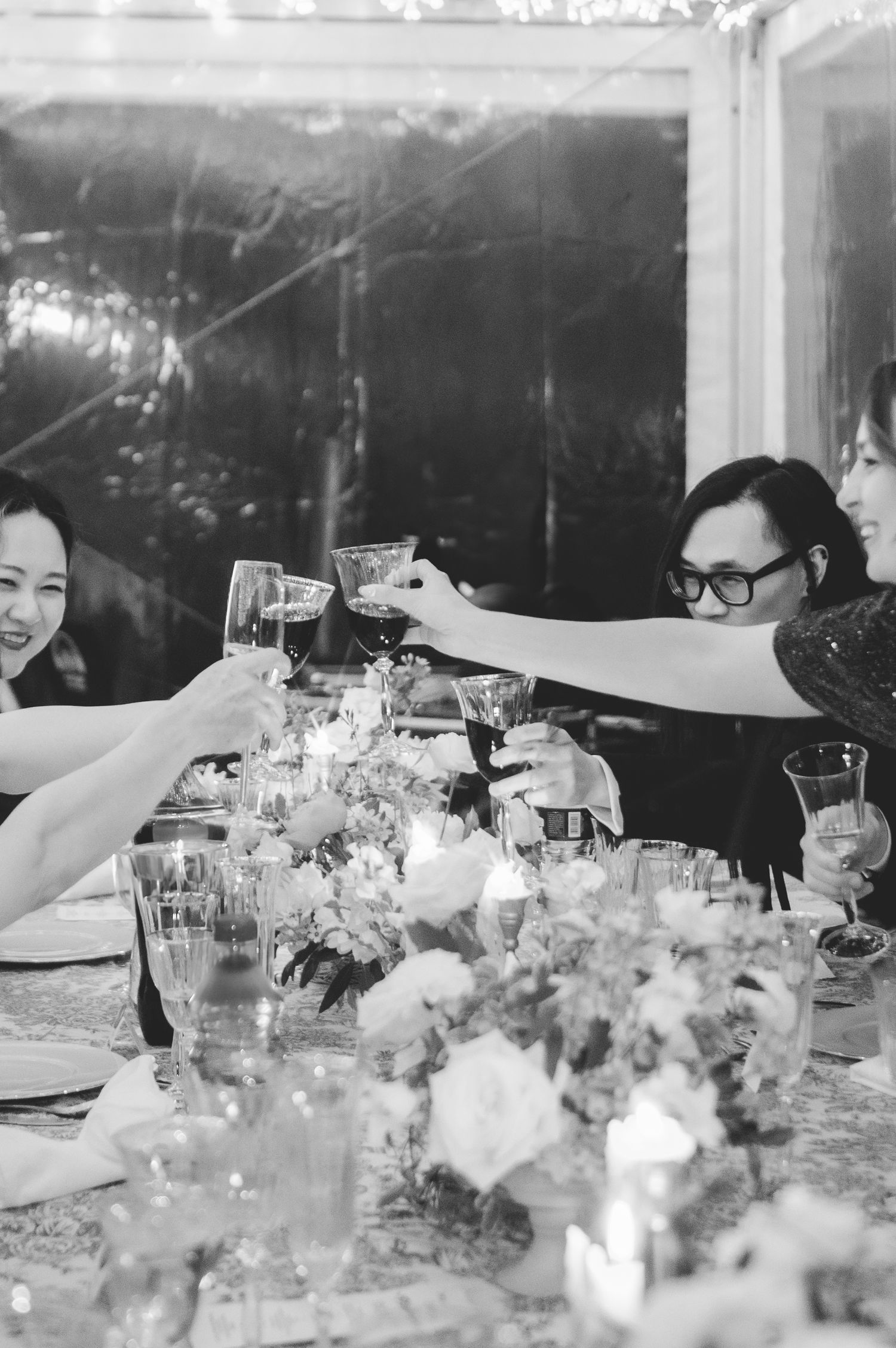 Black and white photo of wedding guests raising glasses in a toast at an elegant candlelit reception dinner.