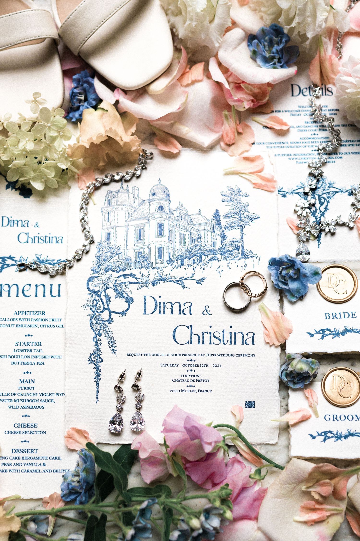 Wedding stationery flat lay featuring blue illustrated invitation suite, rings, earrings, and floral details.