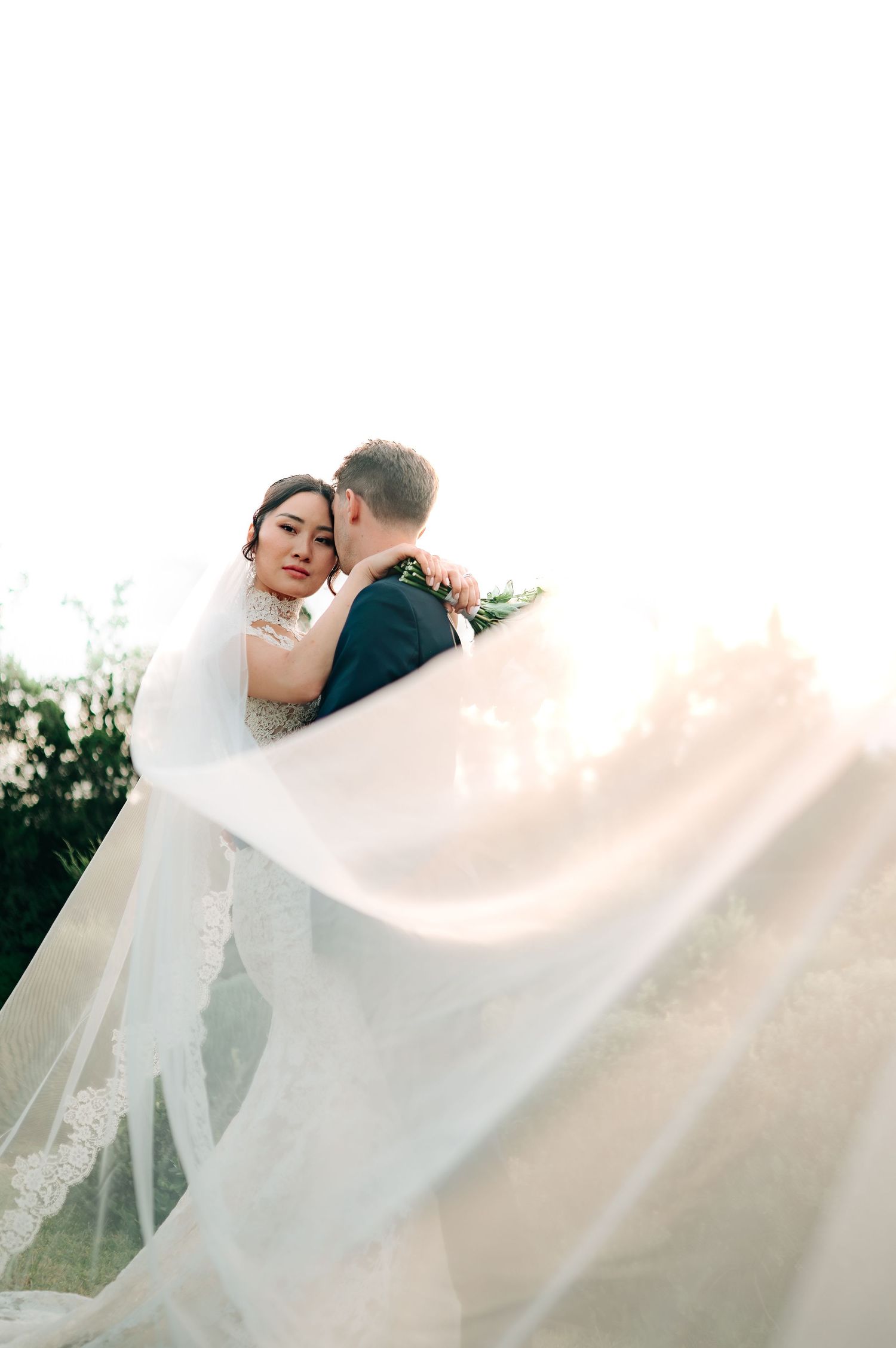 Bride in lace gown with flowing veil embracing groom in sunlit garden, looking softly at the camera.