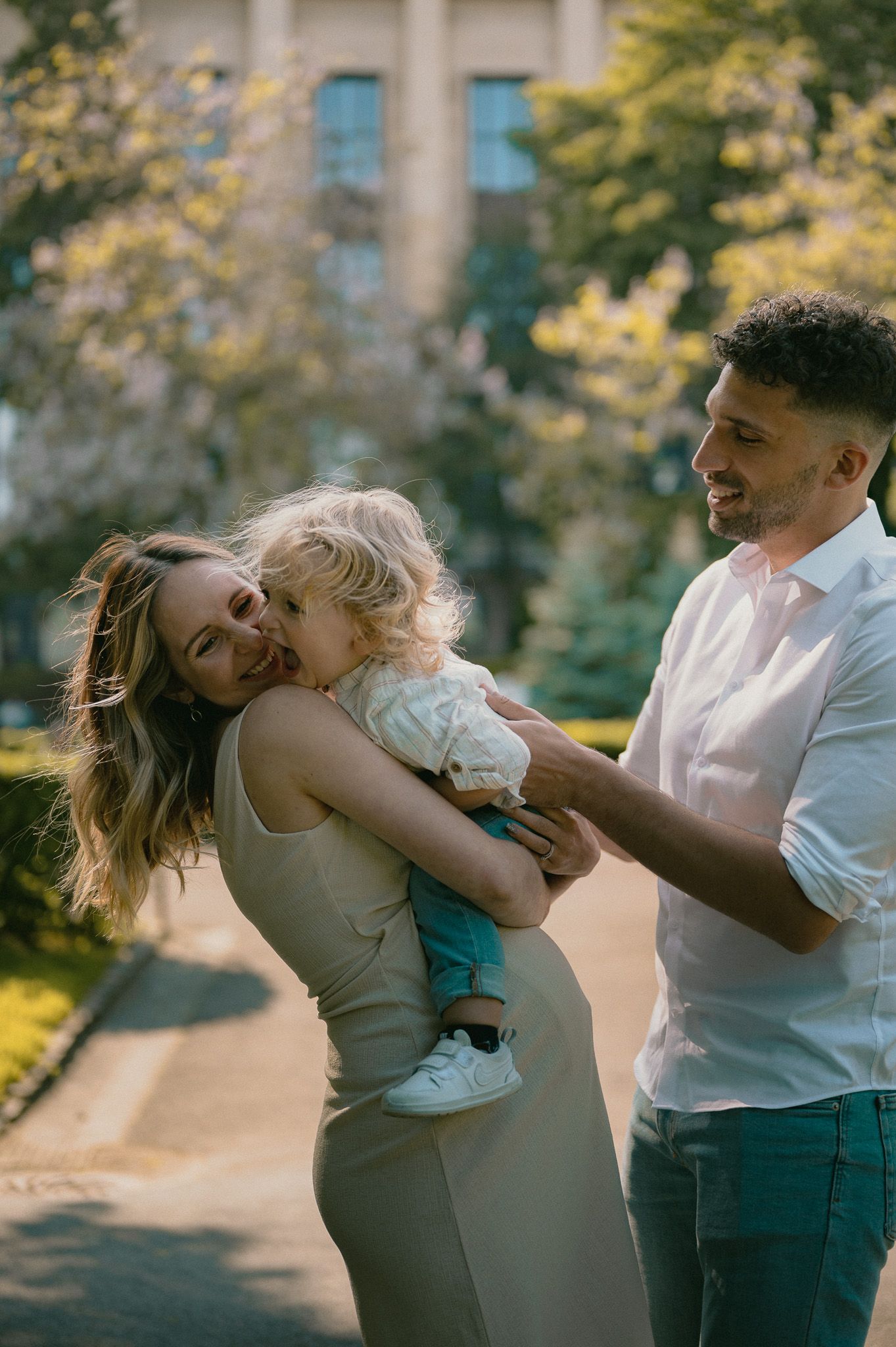 Smiling couple outdoors sharing a joyful moment while holding their curly-haired toddler near blooming trees.