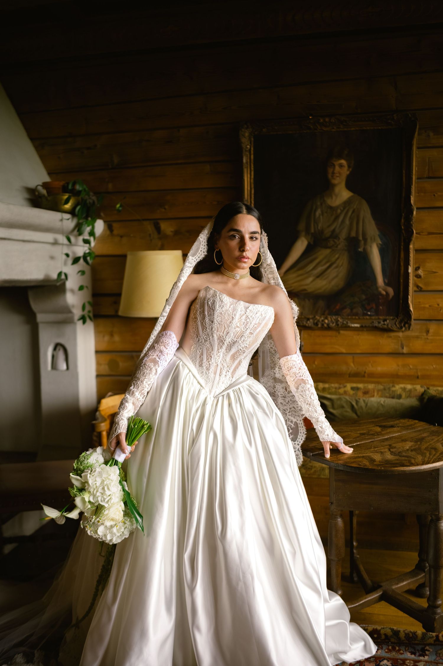 Bride in a stunning strapless ball gown with lace veil holding white bouquet in vintage interior.