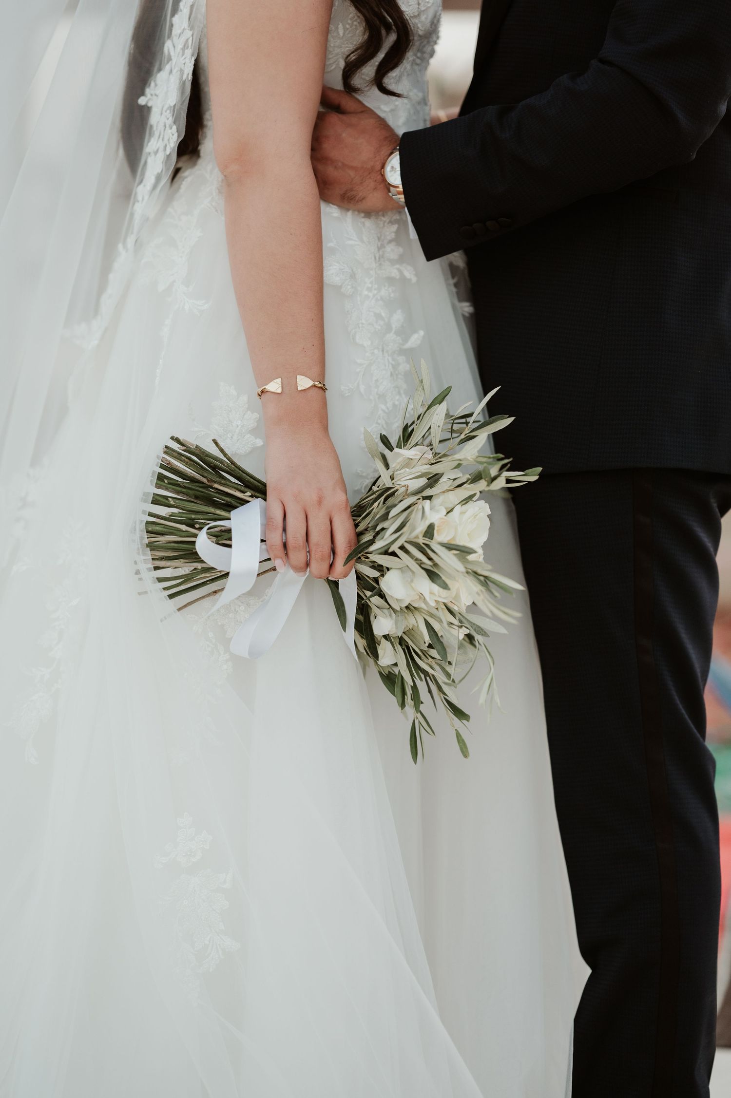 Bride in lace ball gown holds green and white bouquet while groom in black suit embraces her.