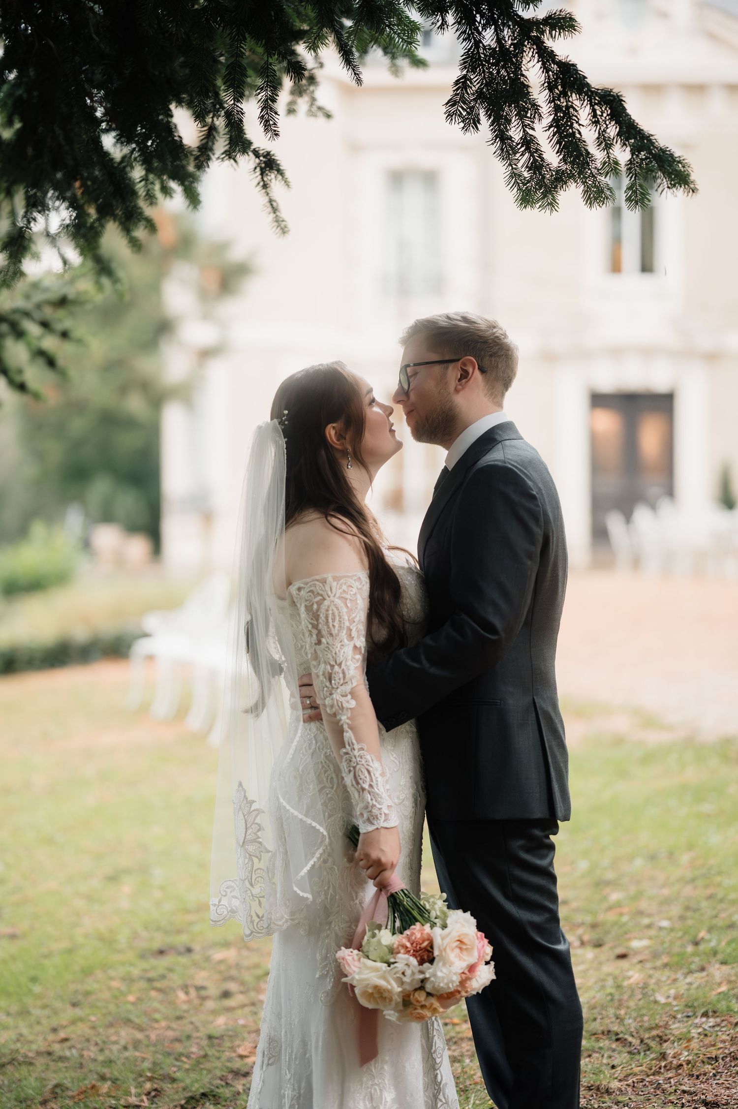 Bride in lace gown and groom in dark suit share romantic moment in chateau garden.
