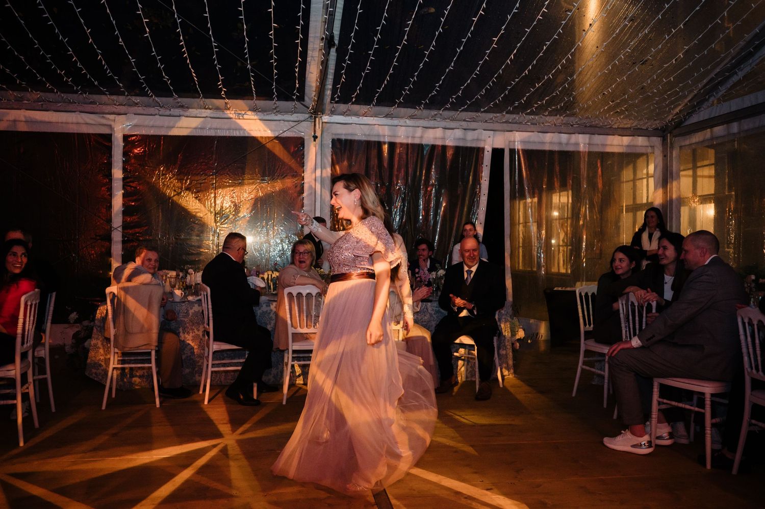 Bride twirls in pink gown on dance floor under twinkling lights at wedding reception.