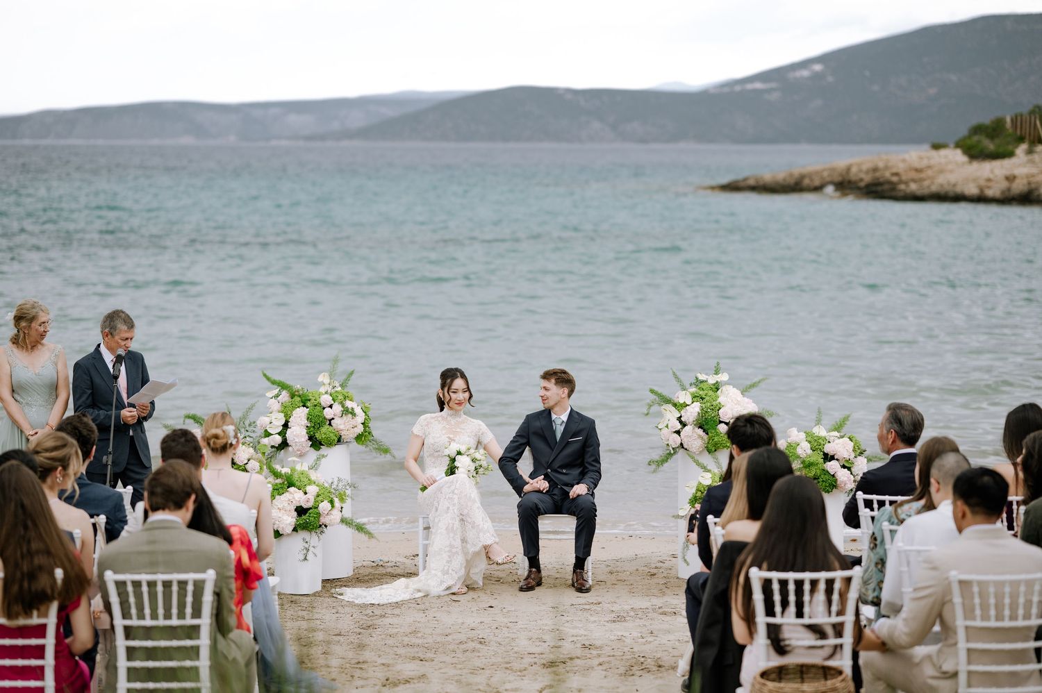 Bride and groom sit during elegant beachside wedding ceremony with sea backdrop.