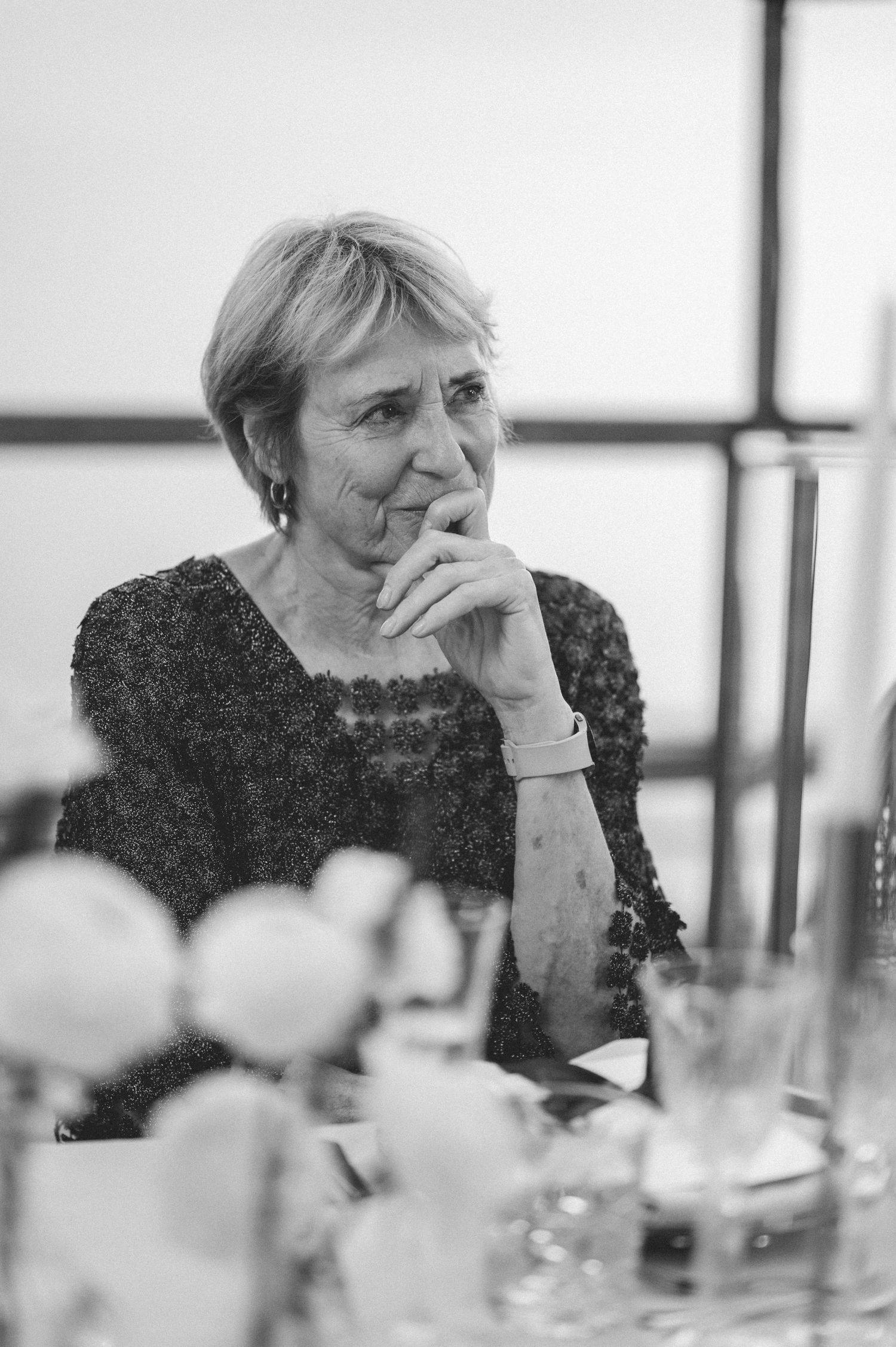 Elegant older woman in embroidered dress seated thoughtfully at wedding reception dinner table.