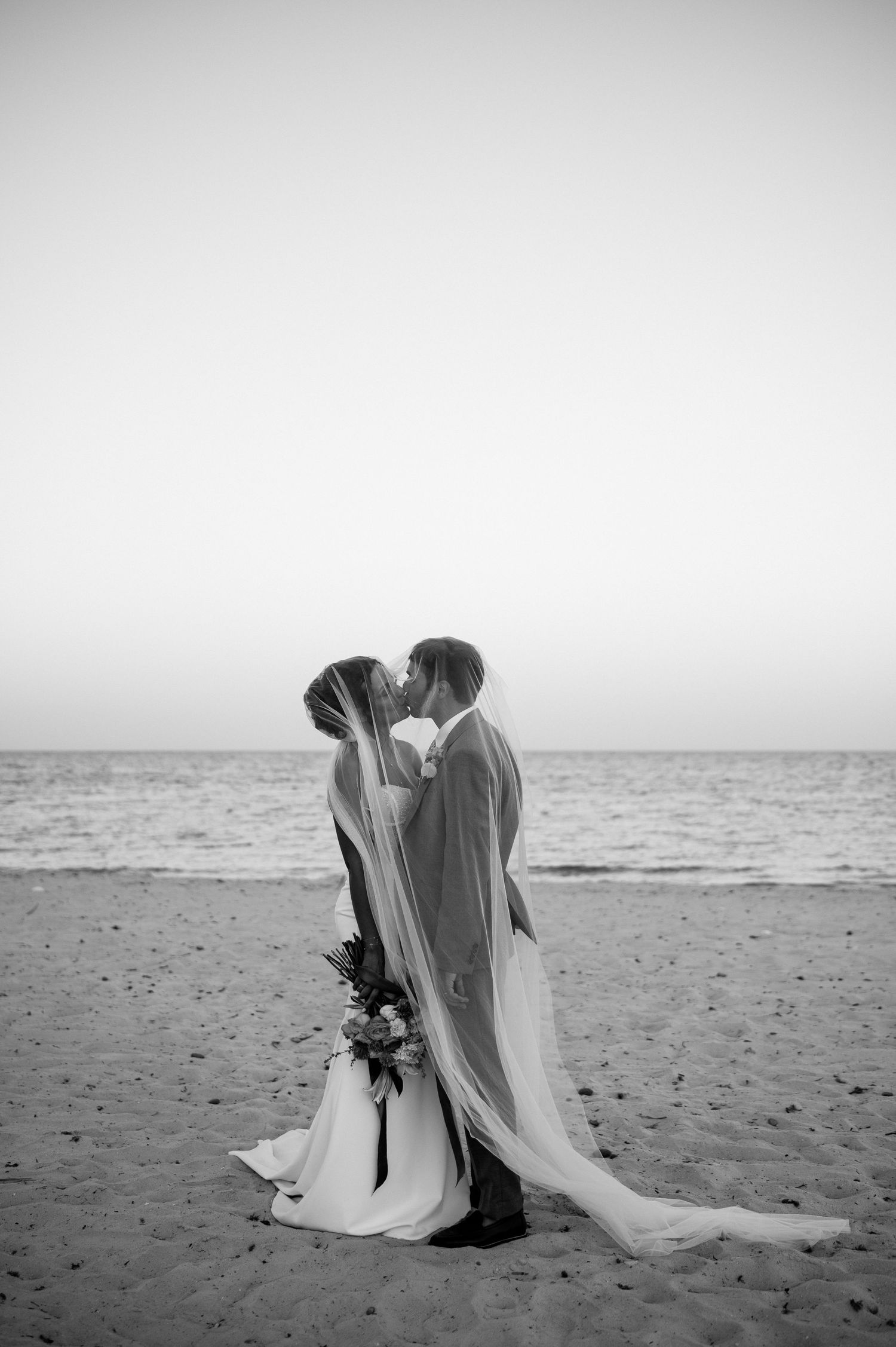 Black and white photo of bride and groom kissing on a beach with ocean backdrop.