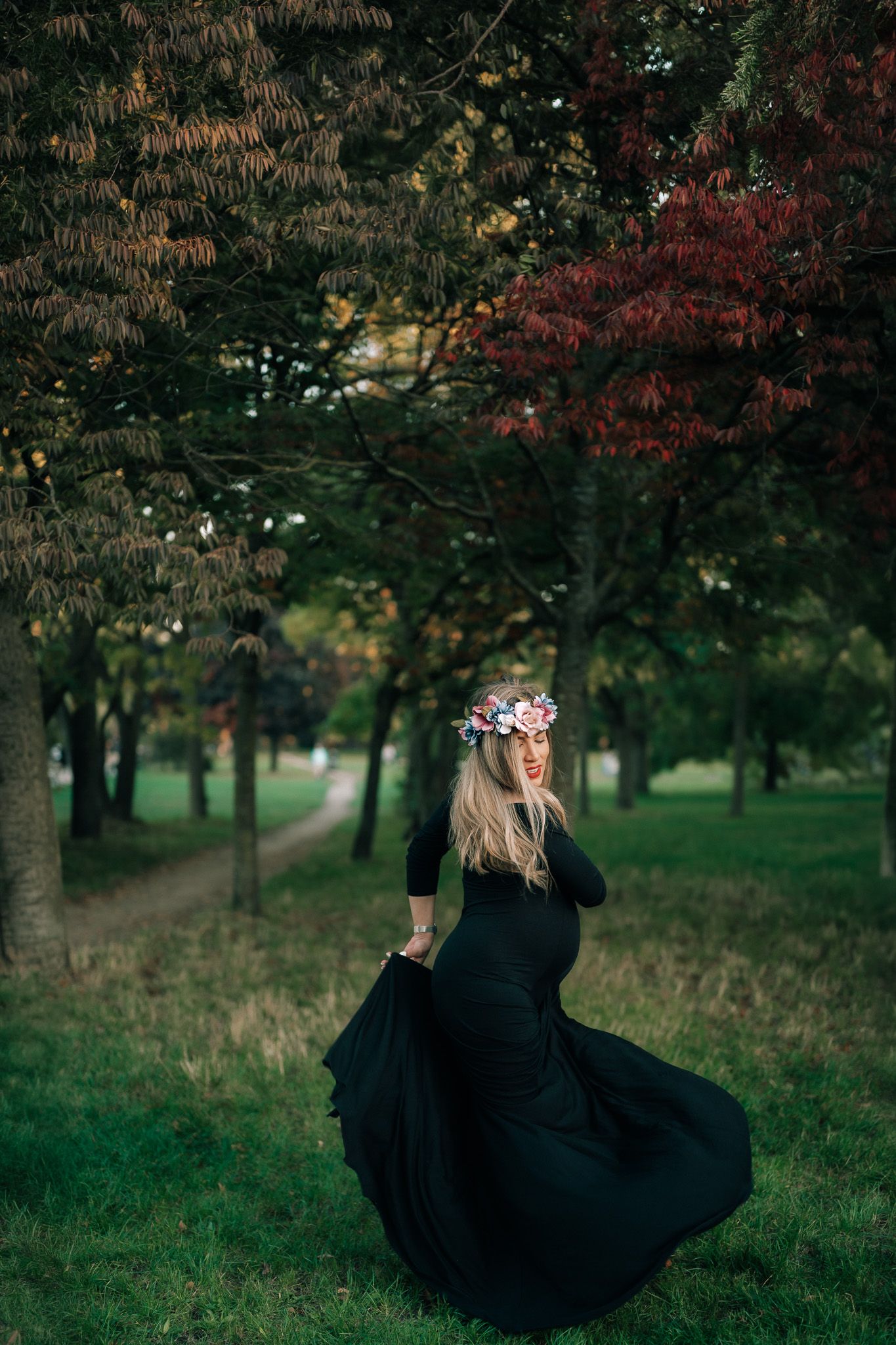 Pregnant woman in black flowing dress and floral crown twirling in a lush green park with colorful autumn trees.