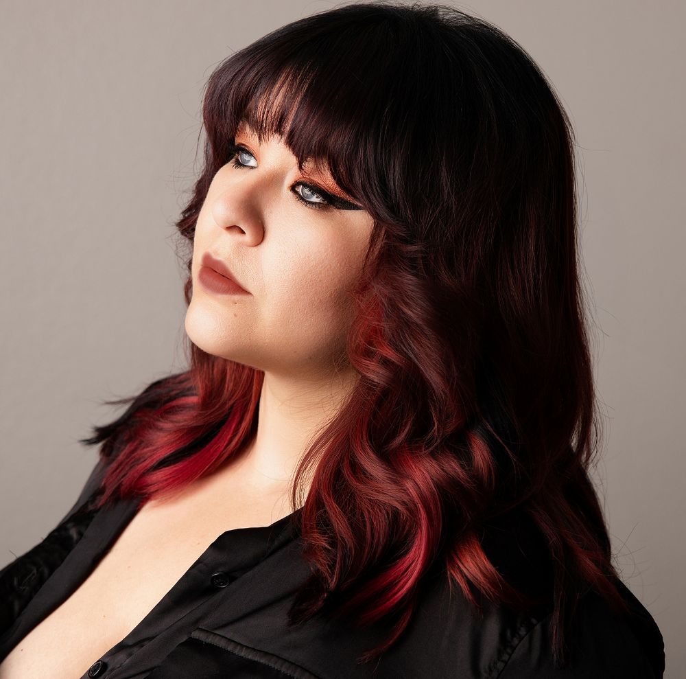 Woman with black and red ombre hair and bangs posing against a gray background in a black top.