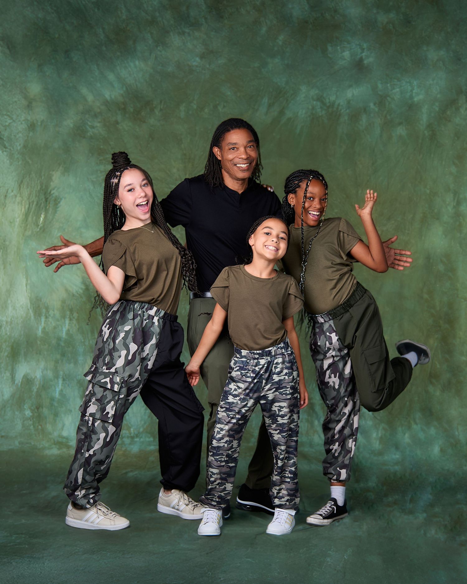 Group of four family members posing in camo pants and olive tops against green backdrop.