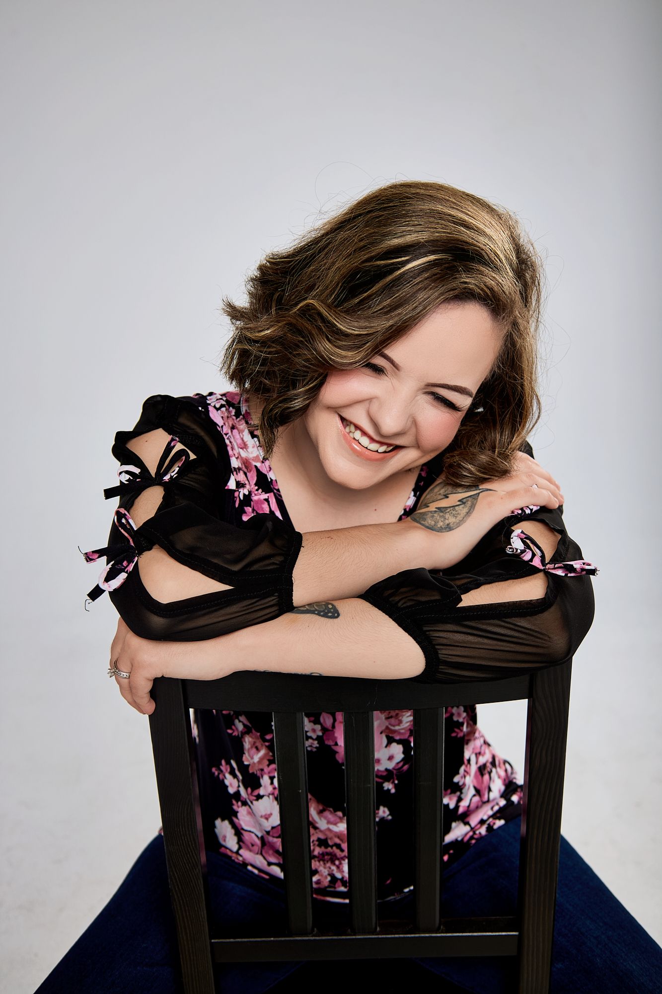 Smiling woman in floral off-shoulder top leaning on black chair in studio portrait.