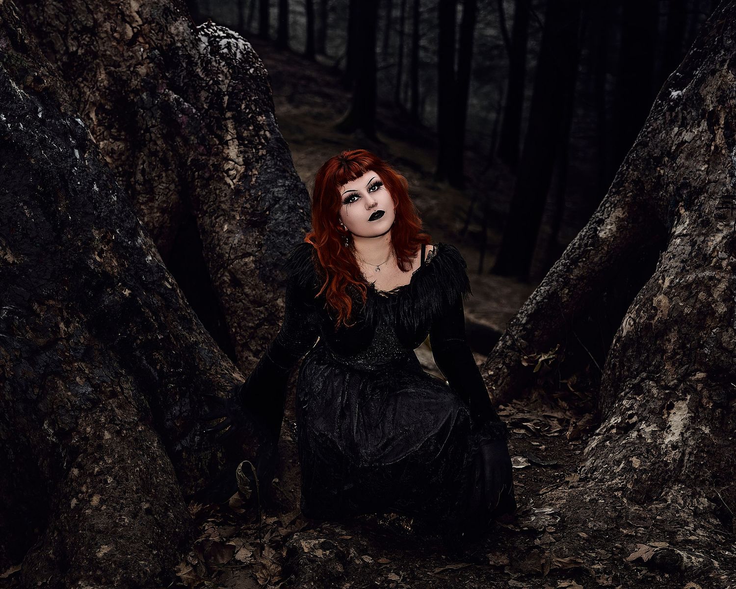 Red-haired woman in black gothic gown kneeling between dark tree roots in forest.
