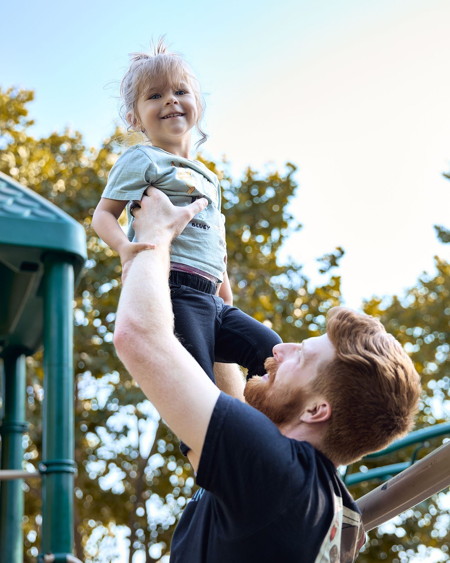 A red-bearded father lifts his smiling toddler high in the air at a playground surrounded by green trees.