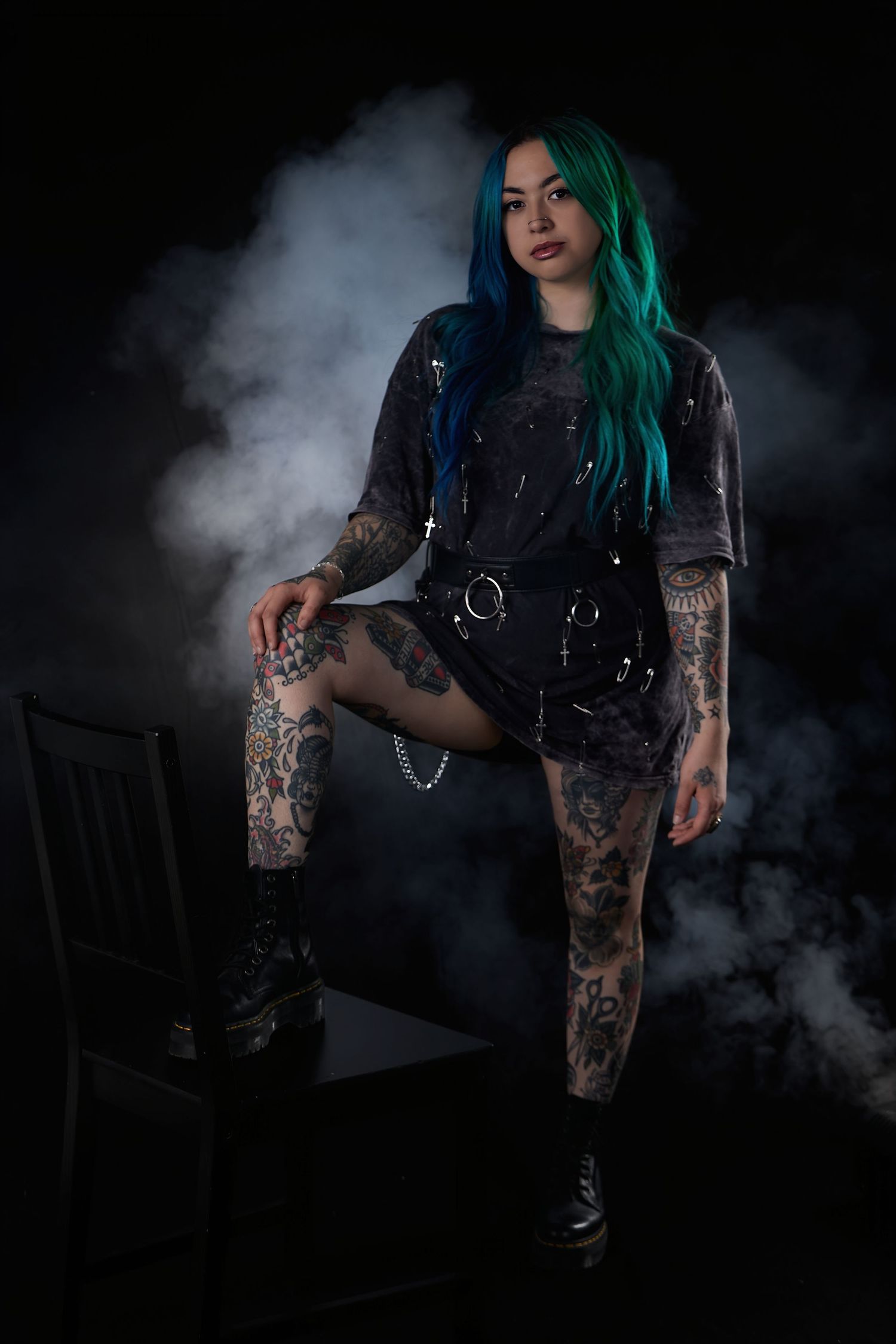 Tattooed woman with teal hair in gothic outfit posing with smoke effects on a dark background.