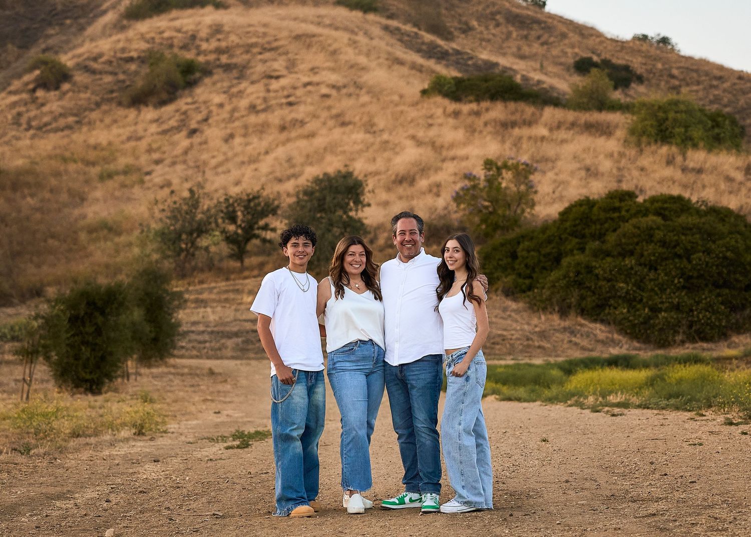 Family of four smiling outdoors on a dirt path with golden hills in the background.