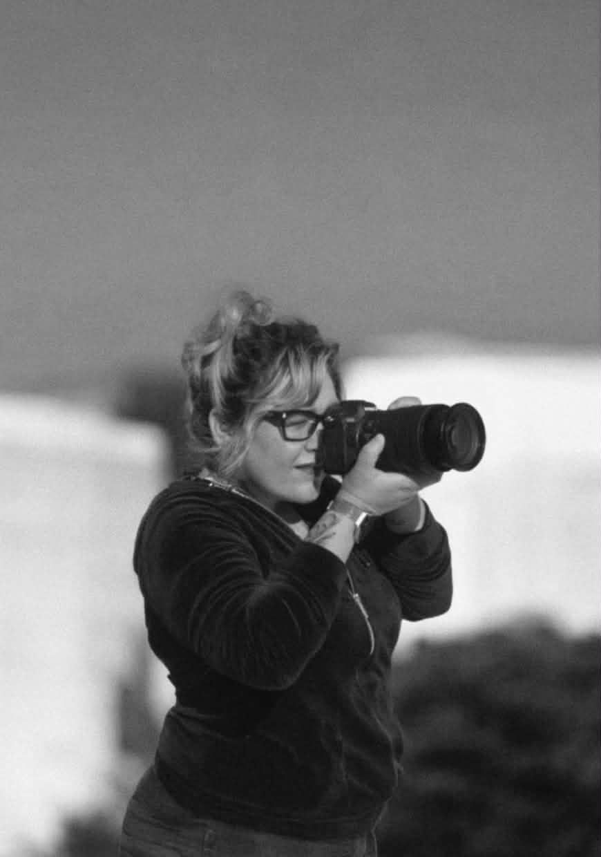 Black and white portrait of woman with glasses photographing with DSLR camera outdoors.
