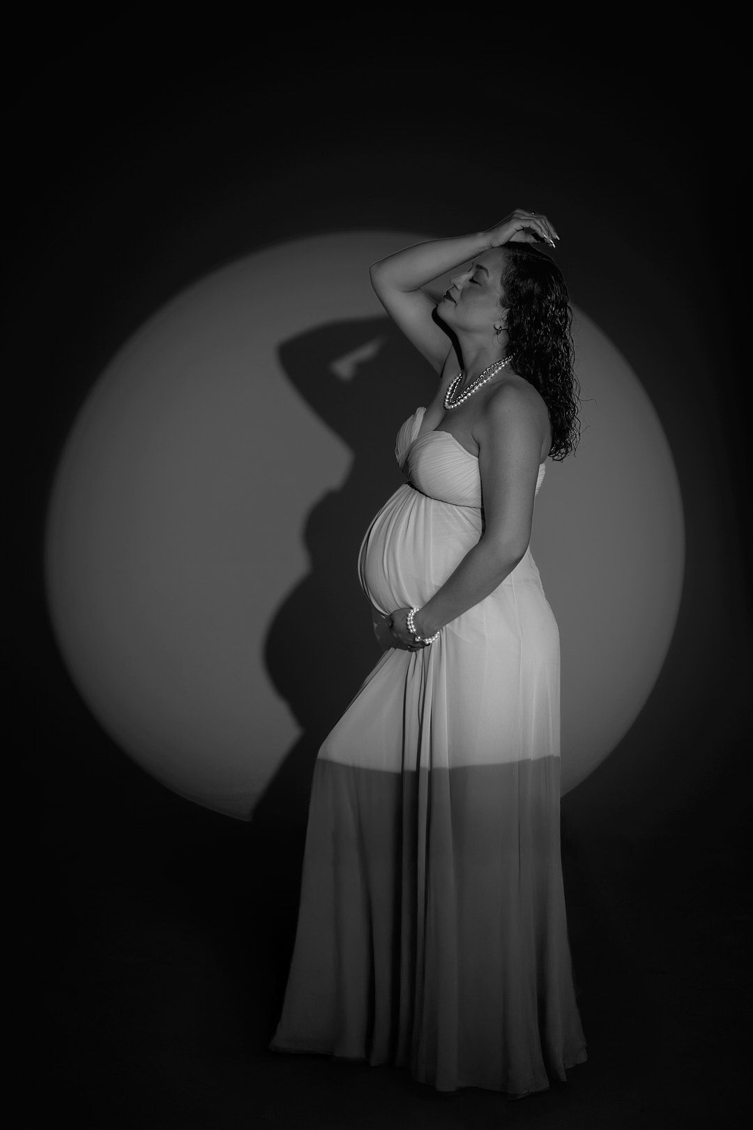 Black and white maternity photo of pregnant woman in flowing white gown with spotlight.