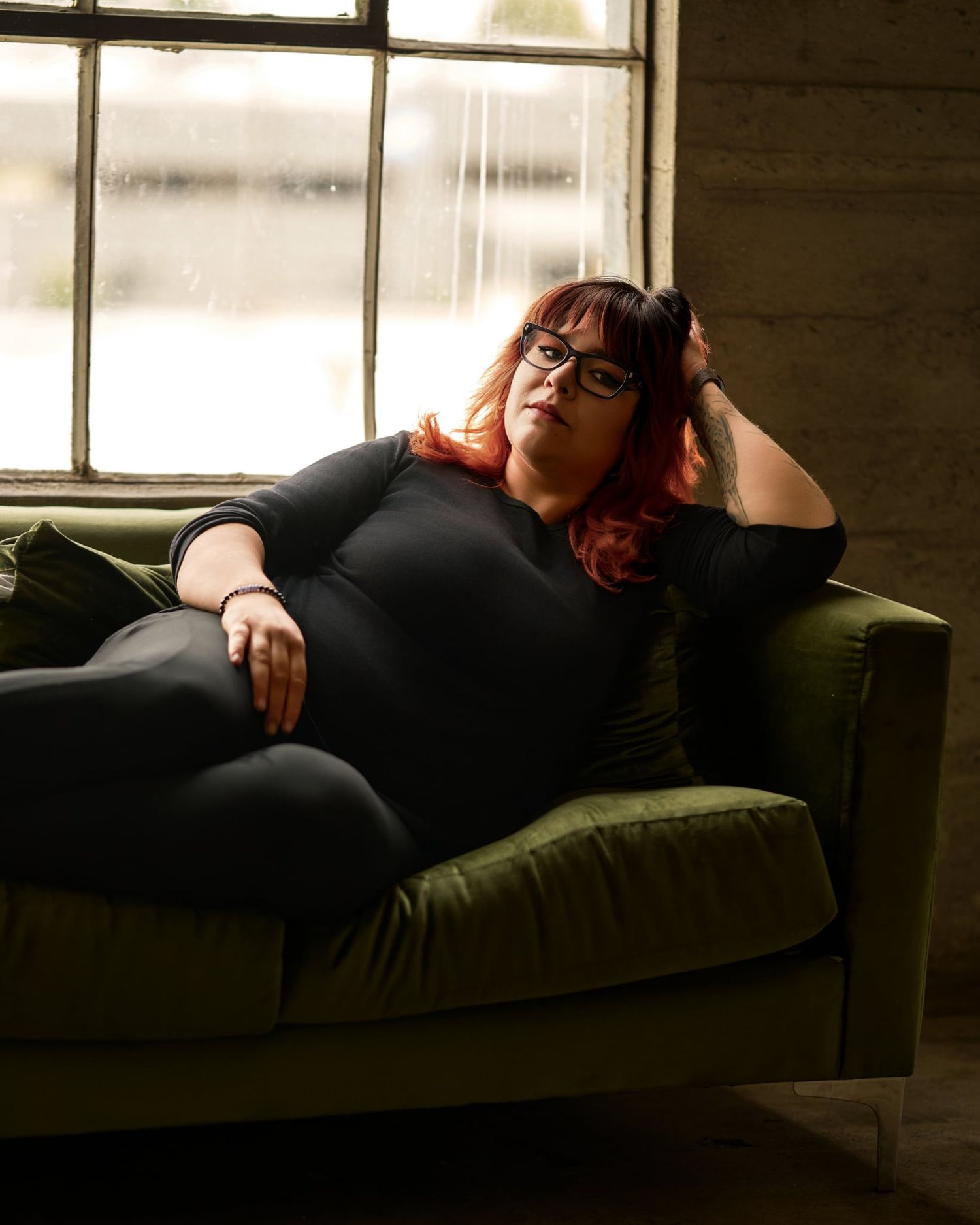Plus-size woman with red hair and glasses relaxing on a green velvet sofa near a rainy window.