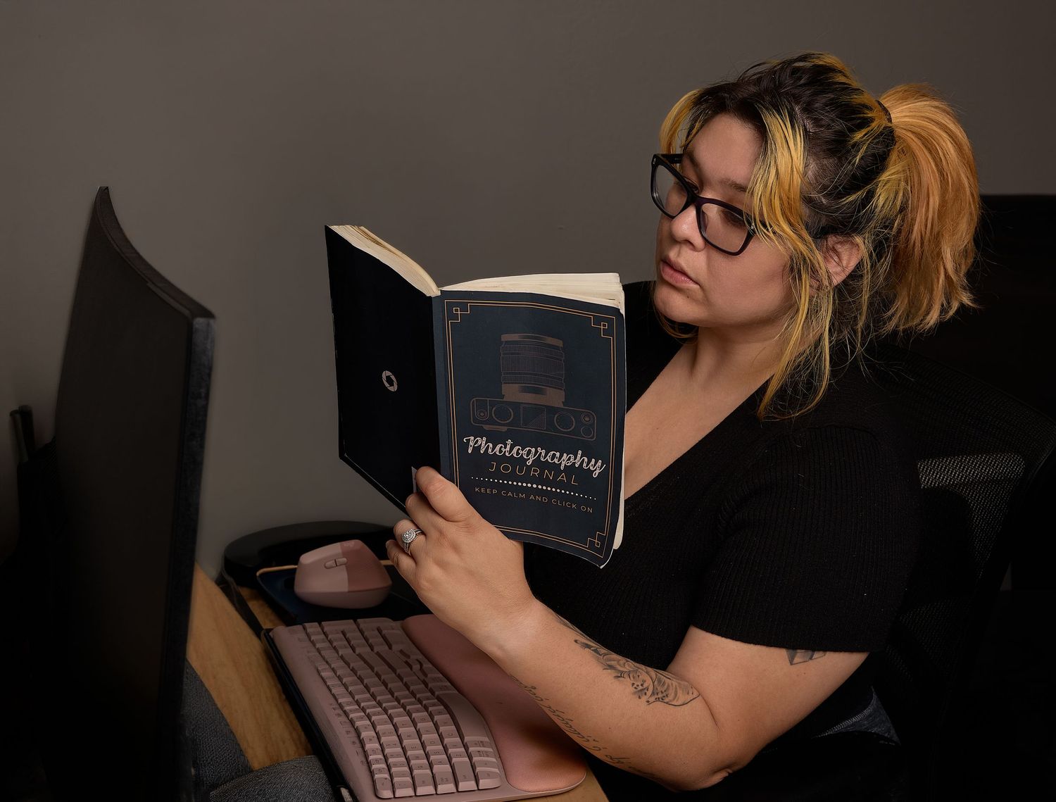 Woman in black top reading a photography book beside a pink keyboard in dim light.