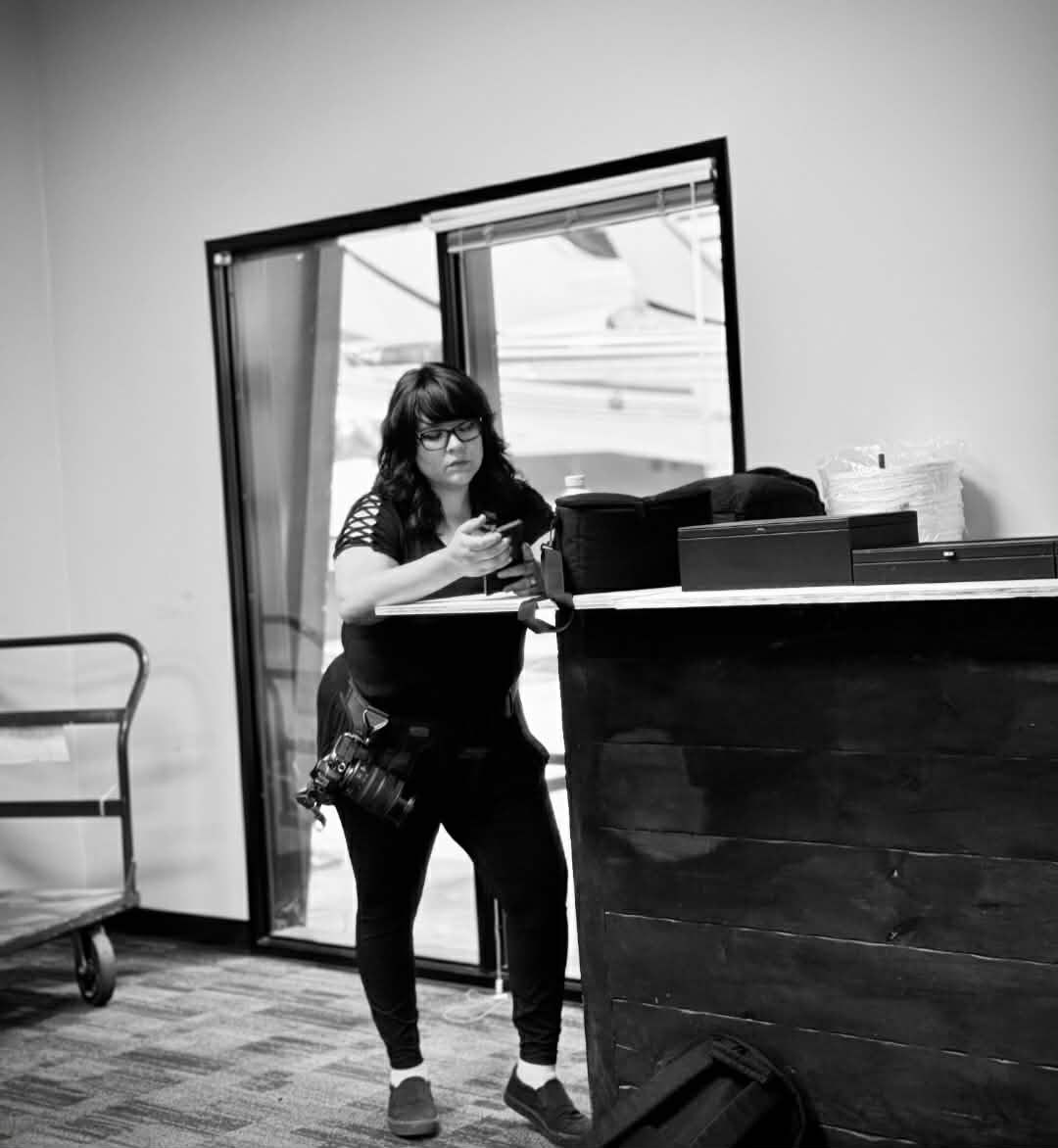 Black and white photo of woman with camera bag working near wooden desk indoors.