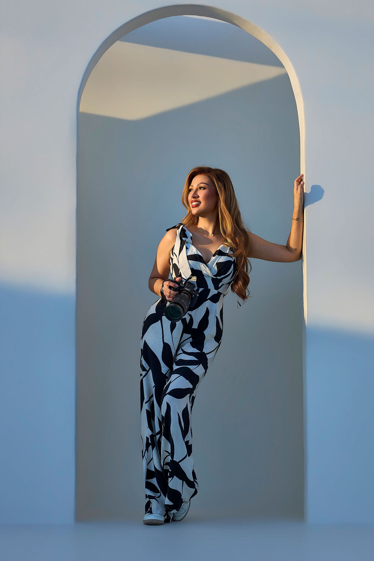 Woman in floral jumpsuit holding a camera standing in an arched doorway.