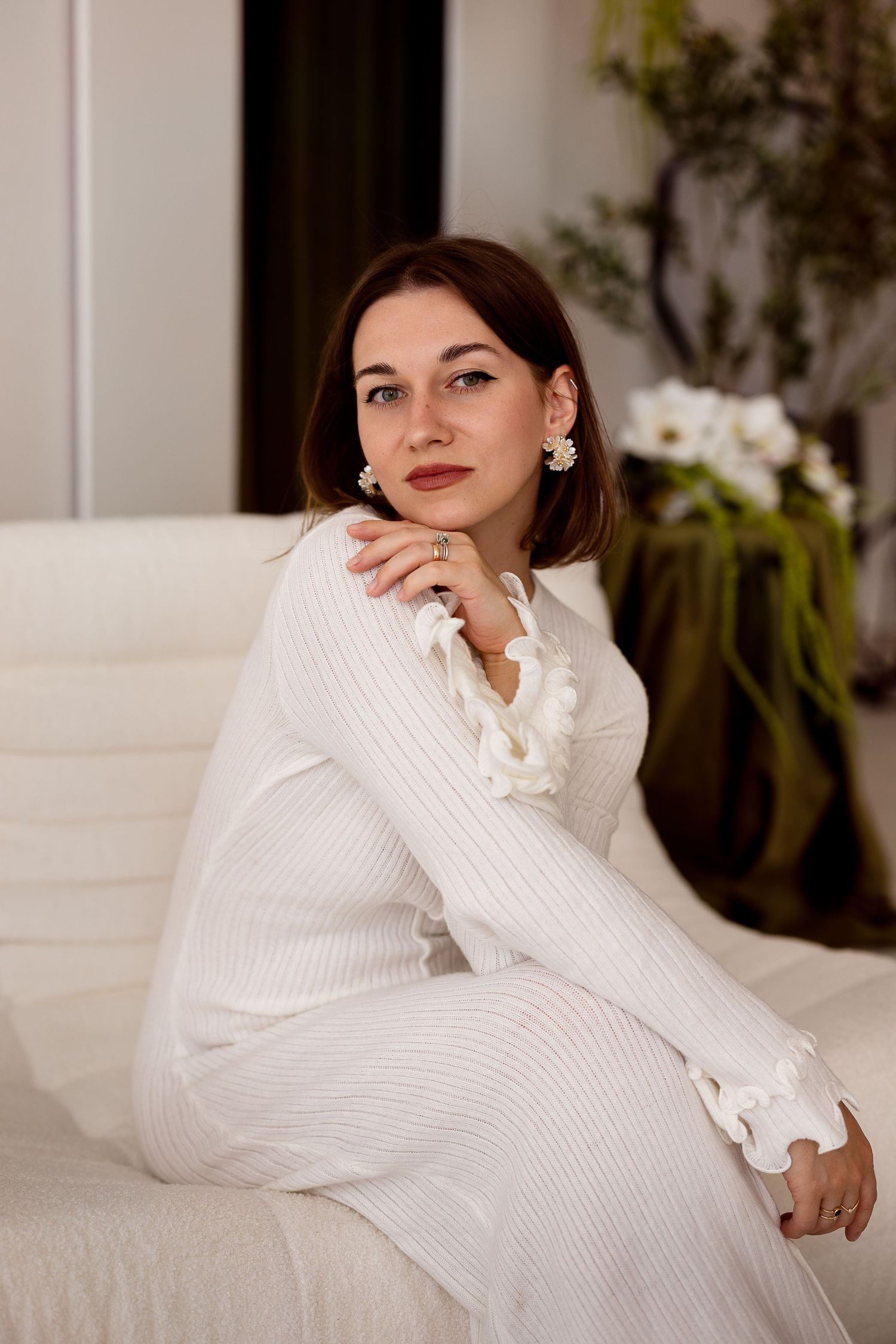 An elegant brunette woman in a white ribbed dress sits gracefully on a cream sofa with pearl earrings.