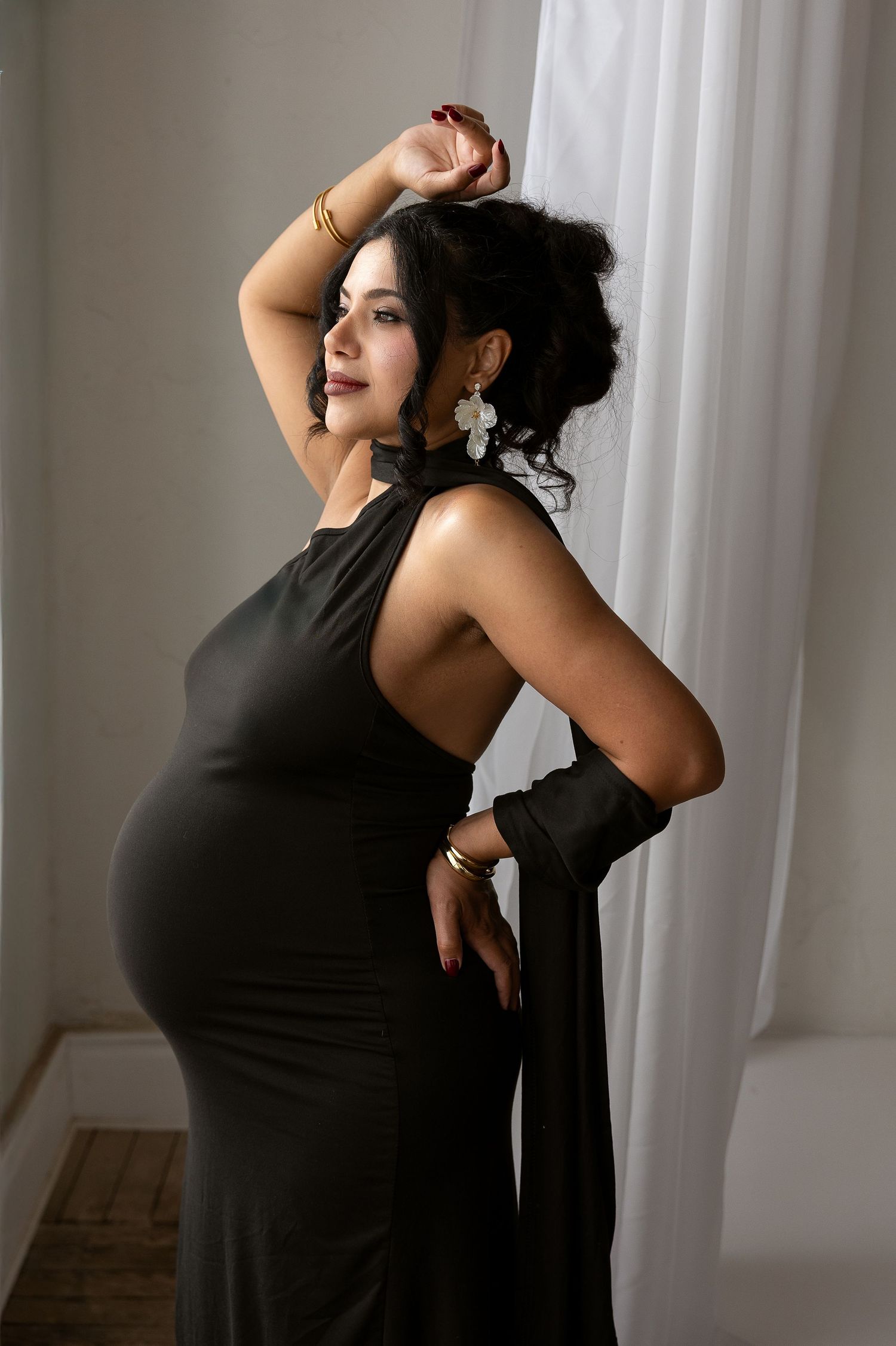 Stylish pregnant woman in black halter dress poses by white curtain, arm raised, wearing gold bangles and drop earrings.
