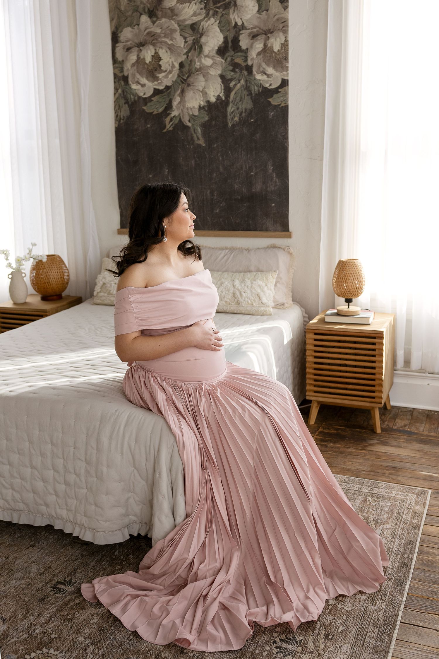 Pregnant woman in blush pink off-shoulder pleated gown sits on bed gazing out bright window.