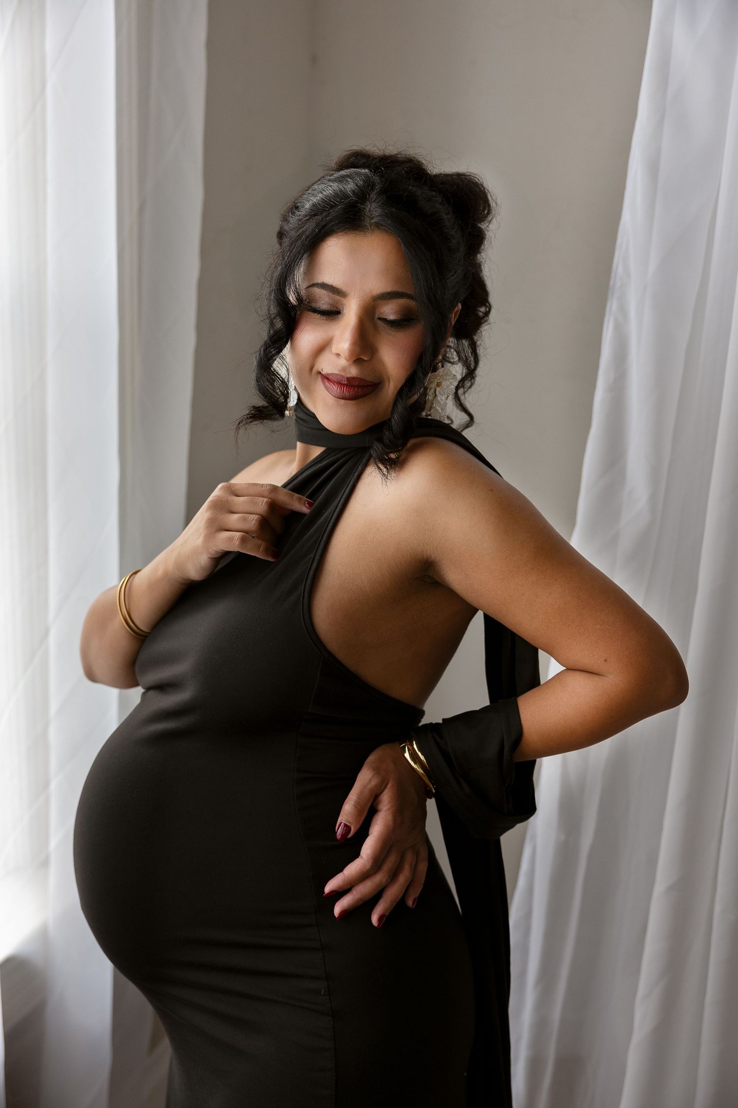 Elegant pregnant woman in dark olive halter dress with gold bangles posing indoors.