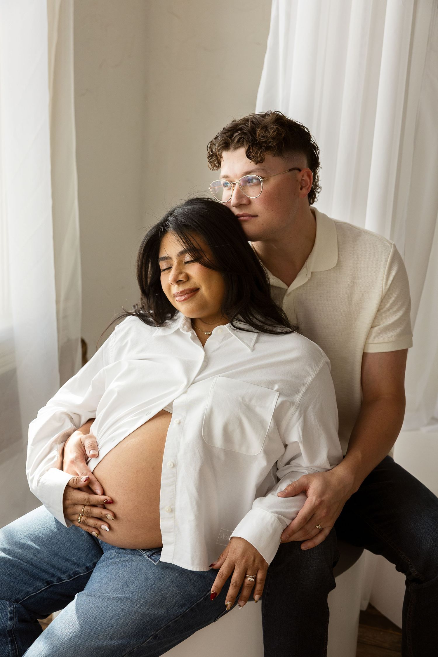 Couple shares tender moment, man rests head on pregnant partner's shoulder near sheer curtains.