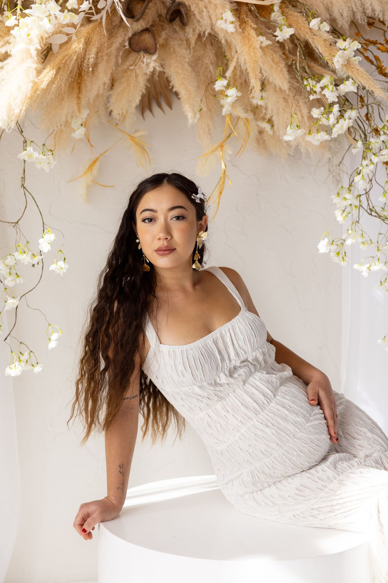 Young woman in white textured dress seated under dried floral arrangement with long wavy hair.