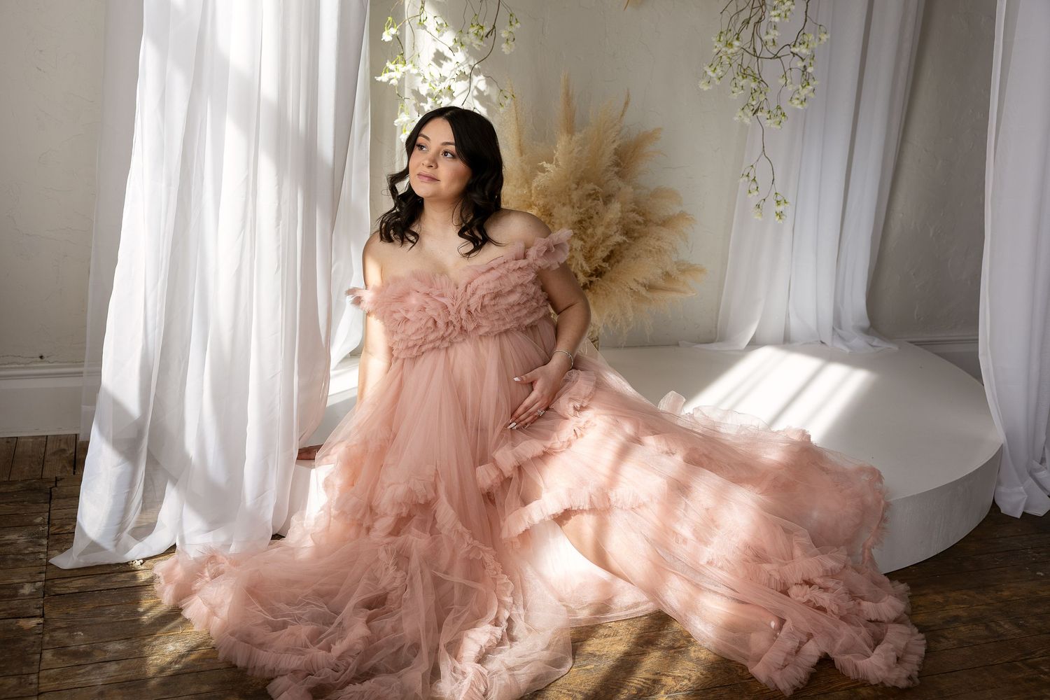 A pregnant woman in a layered blush pink tulle gown sits elegantly in a sunlit, ornate studio setting.