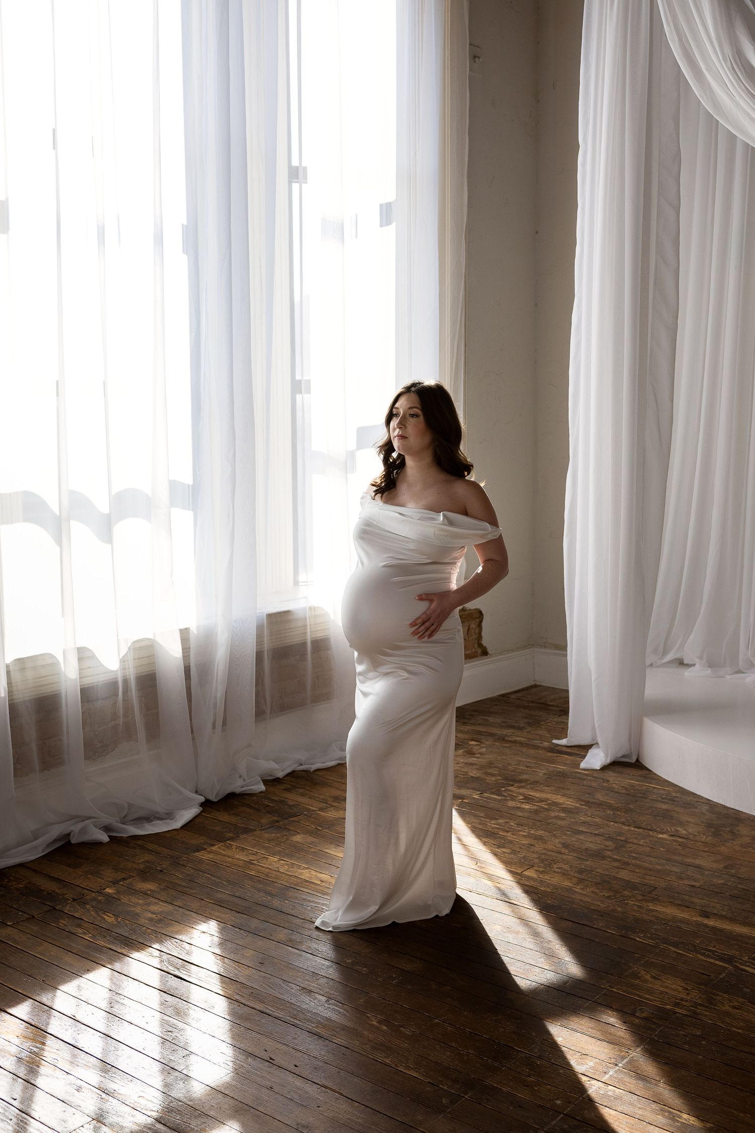 A pregnant woman in a white off-shoulder satin gown stands in sunlit room with sheer white curtains.