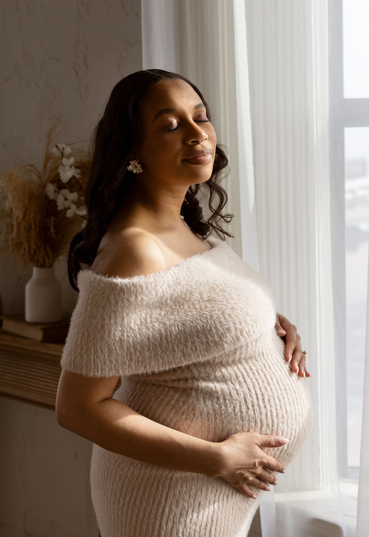 A pregnant Black woman in a fuzzy off-shoulder cream dress cradles her belly near a bright window.