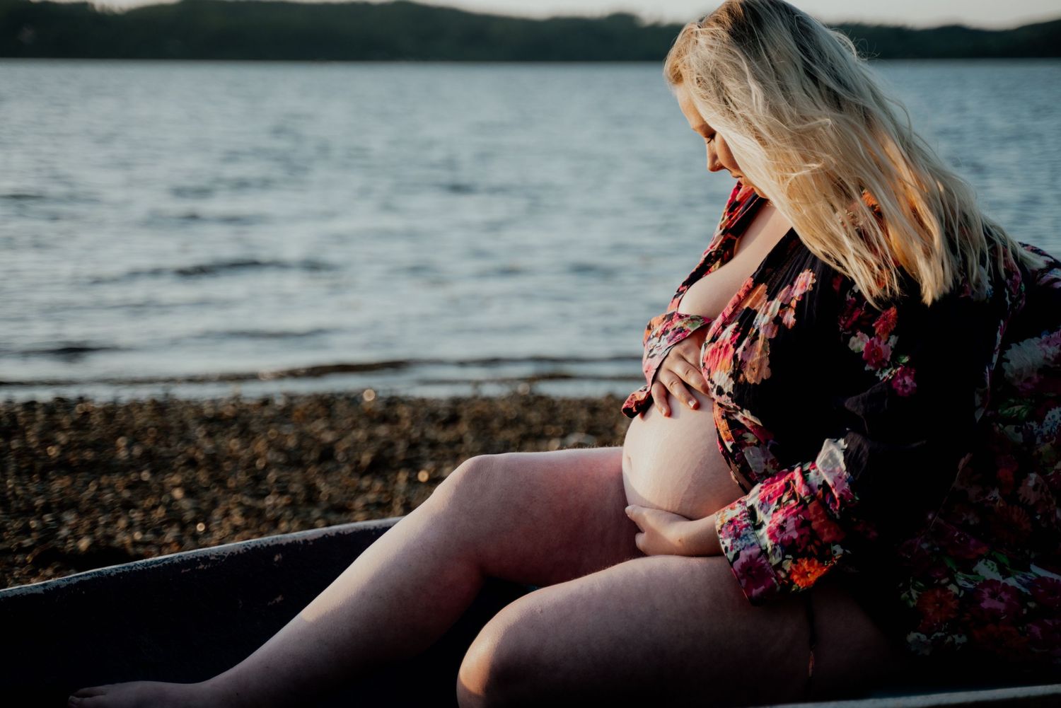 Gravid kvinde siddende ved vandet med hænderne omkring sin mave