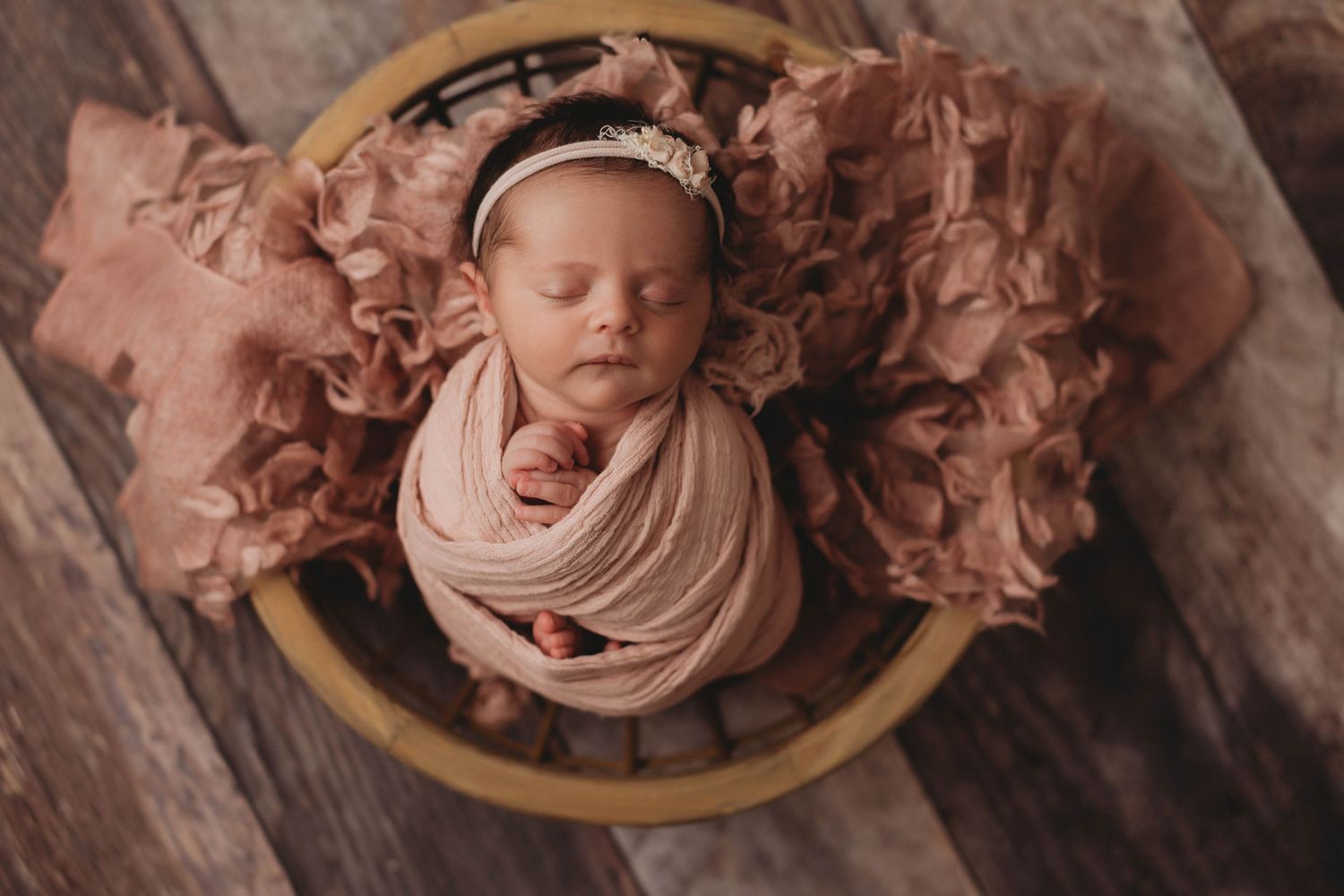 Newborn baby wrapped in soft pink fabric sleeps peacefully in round wooden bowl surrounded by flowing material.