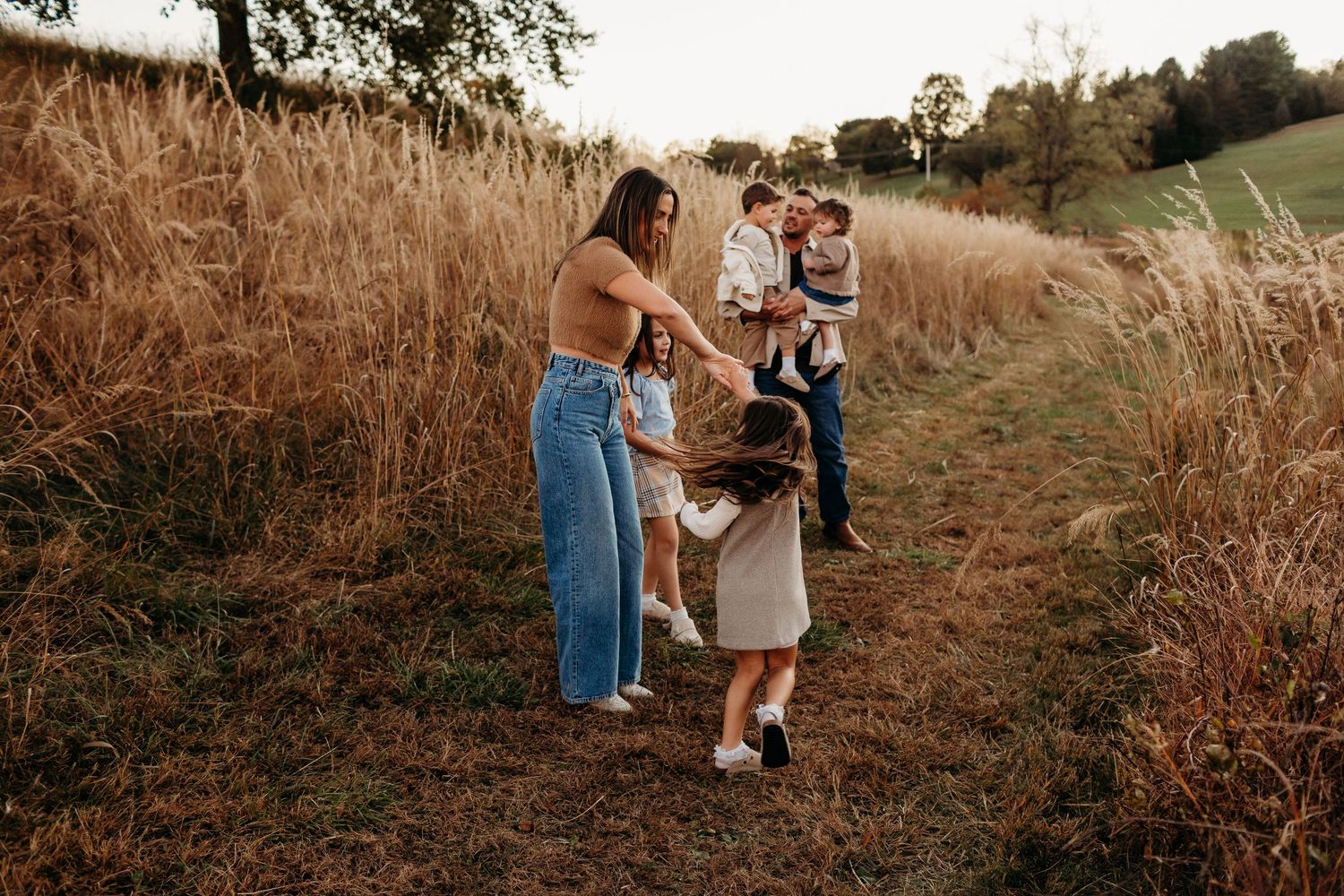 Young family with children playing together on golden grass field path at sunset.
