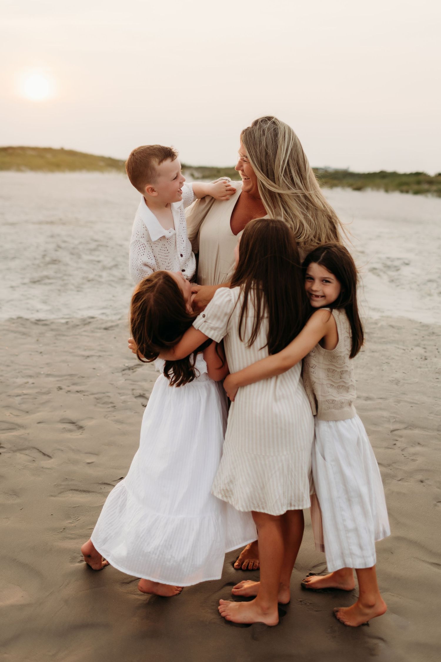 A mother embraces her three children on a sandy beach at sunset, all wearing white outfits in a heartwarming family moment.