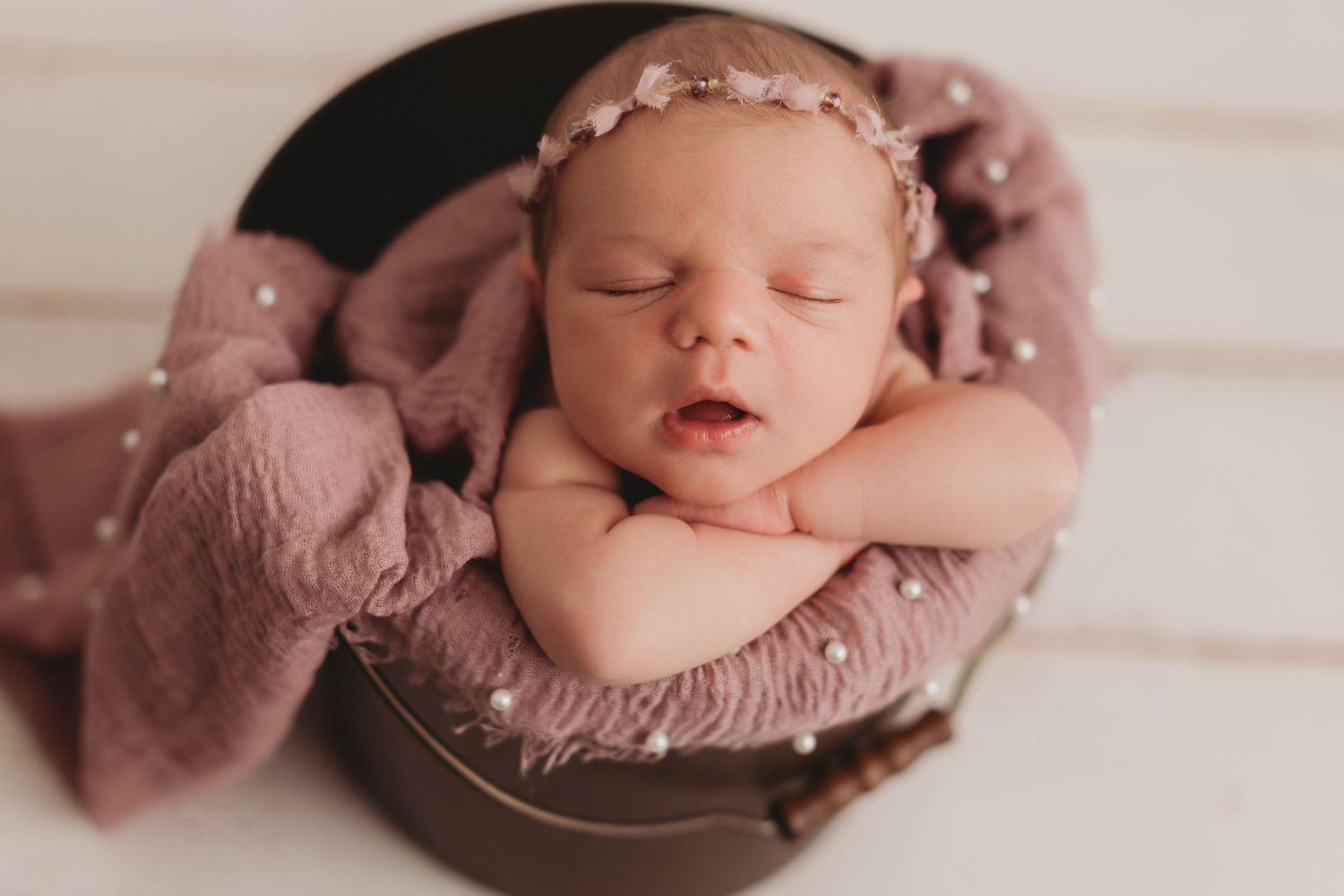 Newborn baby sleeping peacefully in round wooden bowl with dusty pink wrap and delicate headband.