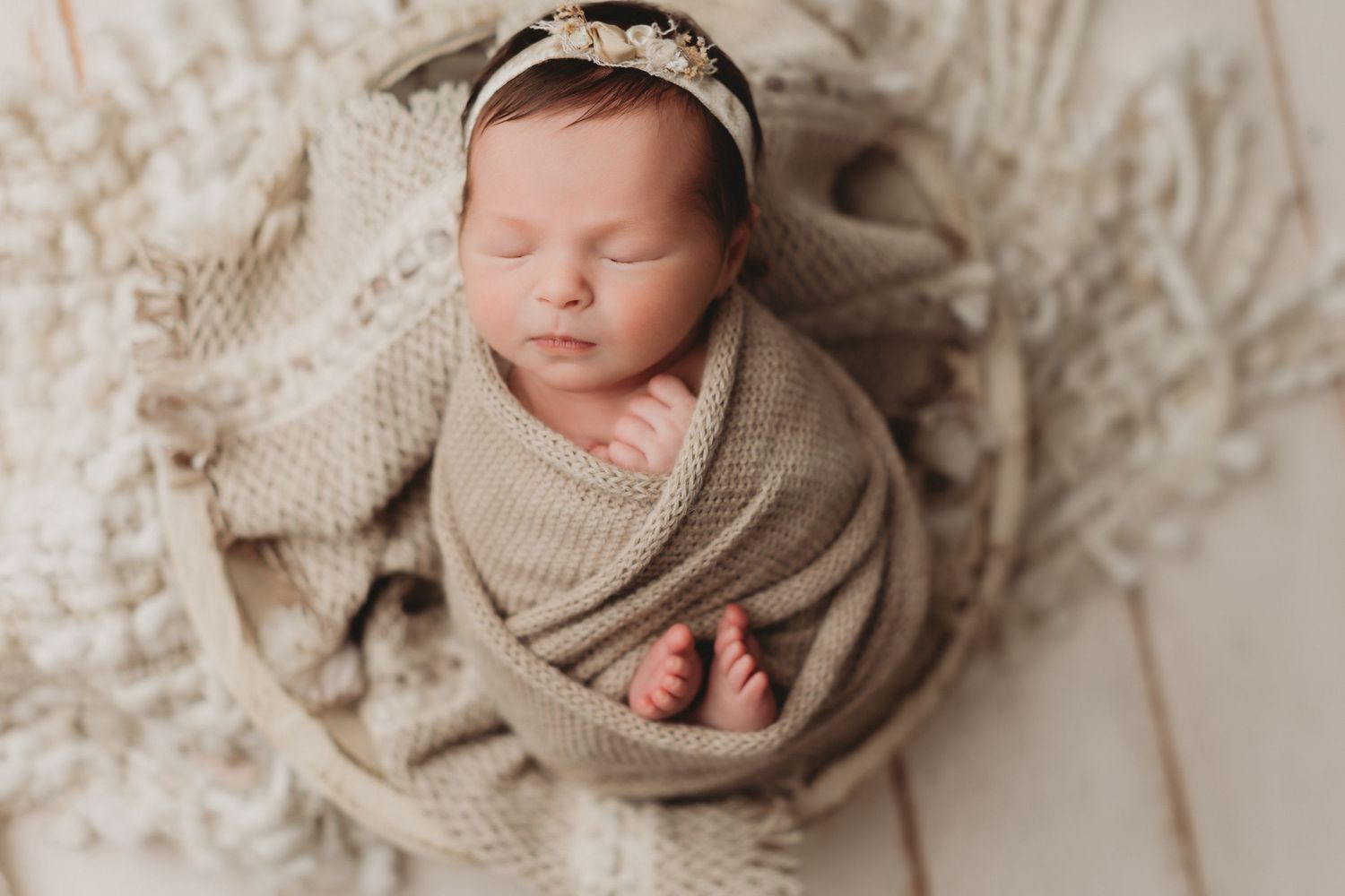 Newborn baby girl swaddled in a beige knit wrap, sleeping peacefully in a round basket with a floral headband.