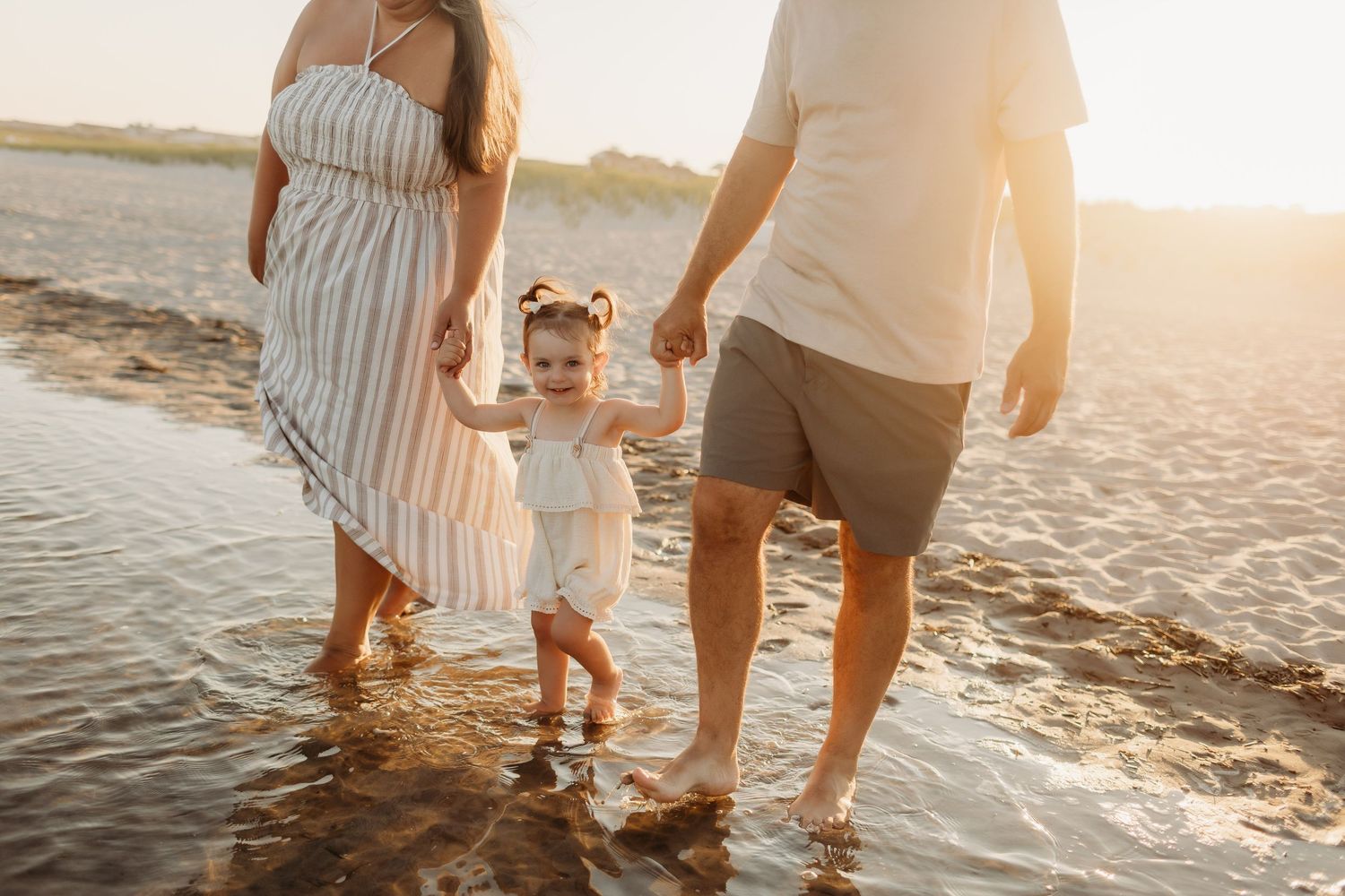 Parents holding toddler girl's hands walking through shallow beach water at golden sunset.