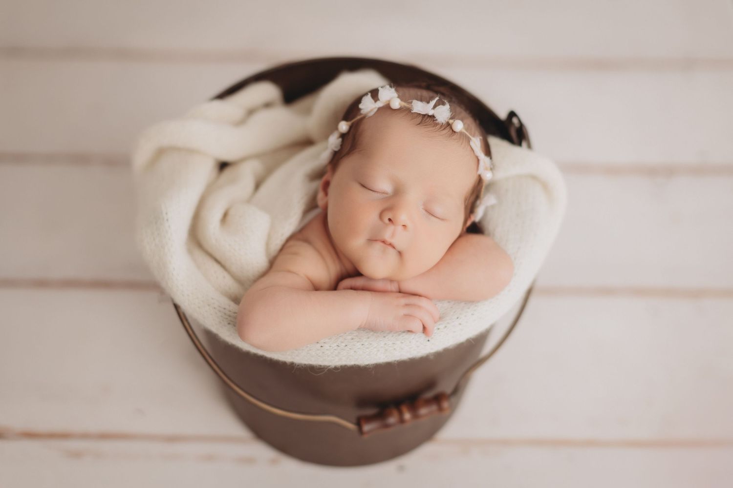 Sleeping newborn baby girl in a rustic bucket with white blanket and floral headband, professional newborn photography.