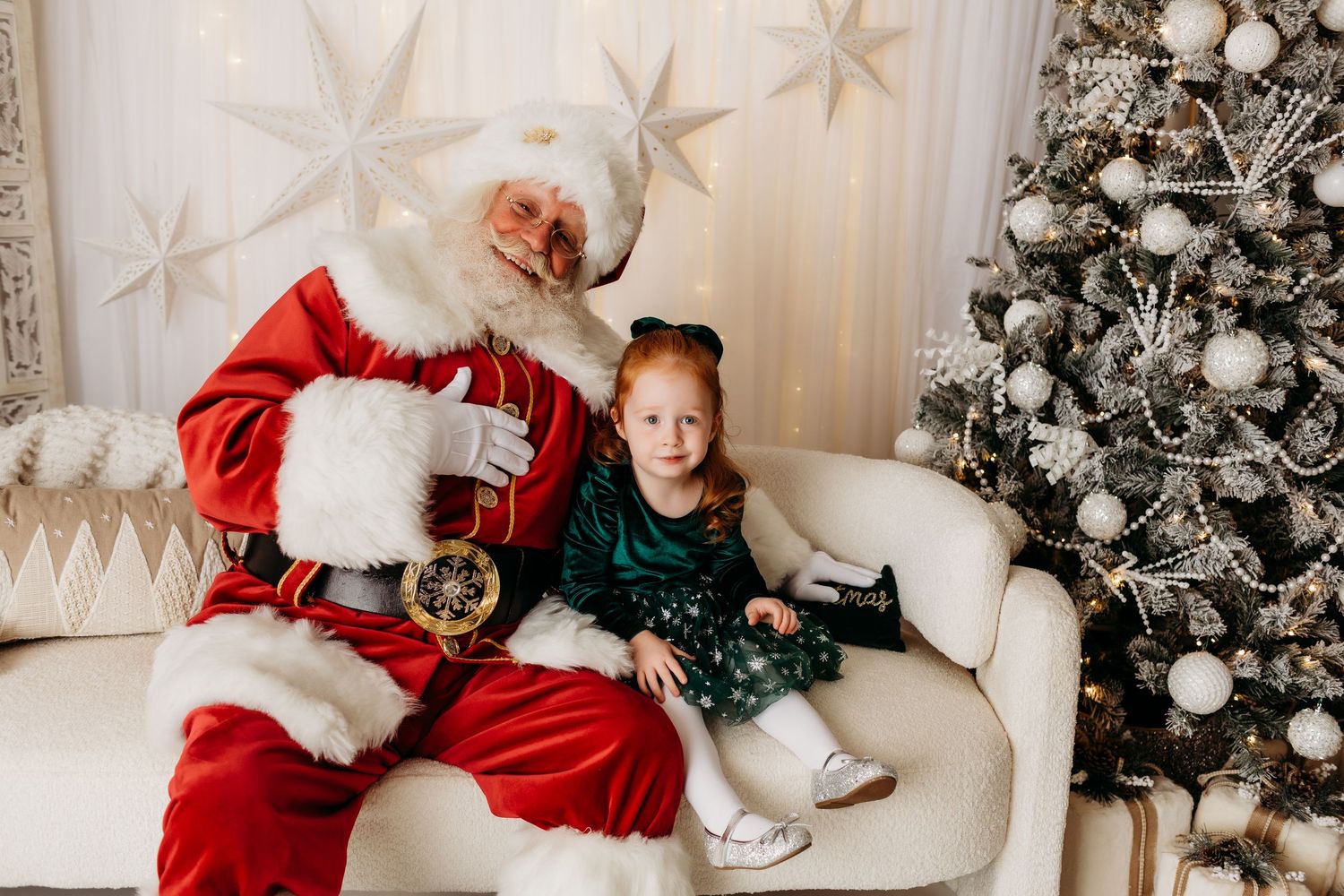 A young girl in a green dress sits smiling with Santa Claus beside a silver and white Christmas tree.