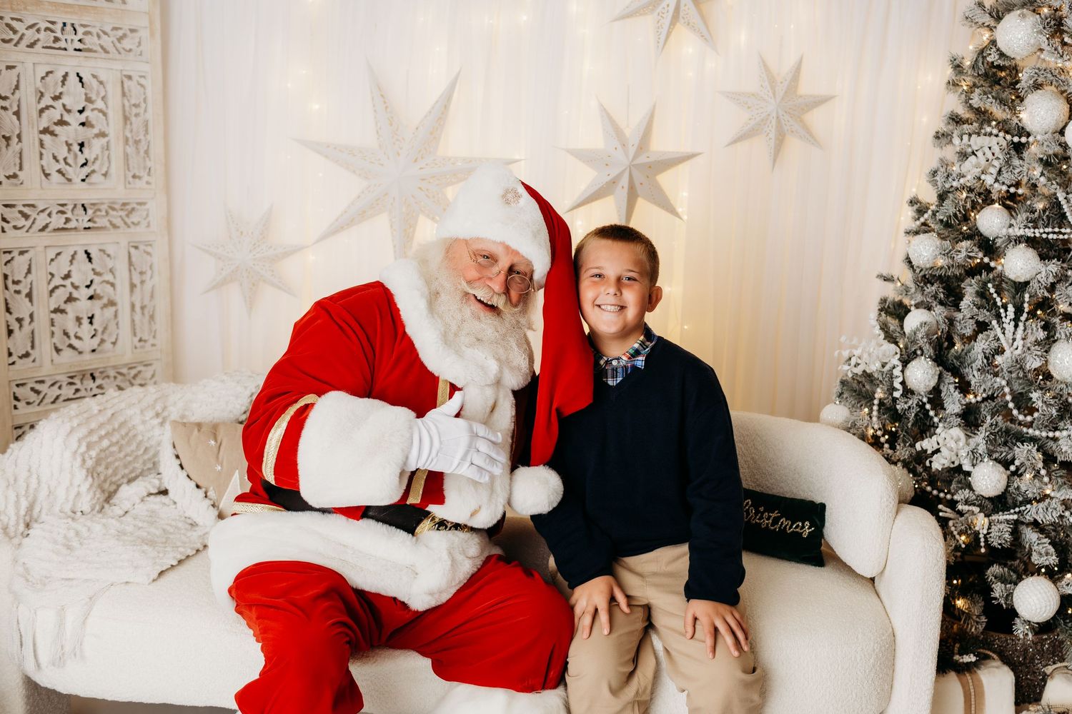A young boy in a navy outfit poses with Santa Claus on a white sofa beside a silver Christmas tree.