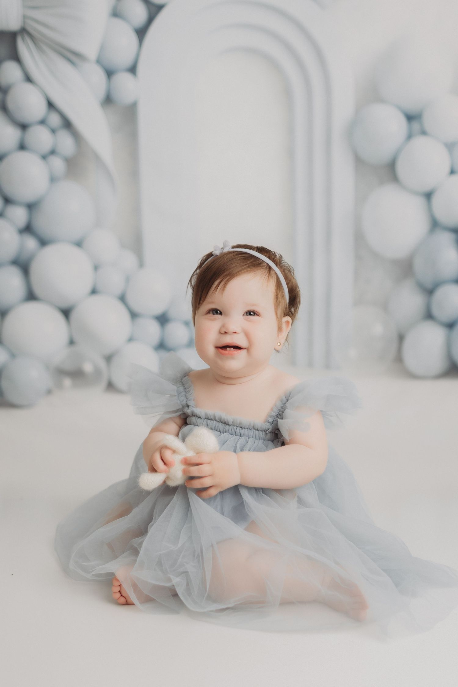 A smiling baby girl in a blue tulle dress sits among blue and white balloon decorations.