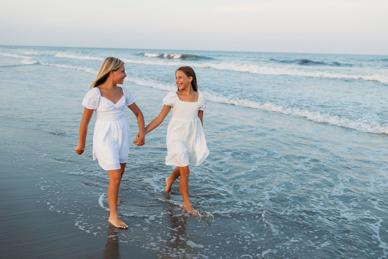 Two people in white dresses hold hands while walking along the ocean shoreline at sunset.