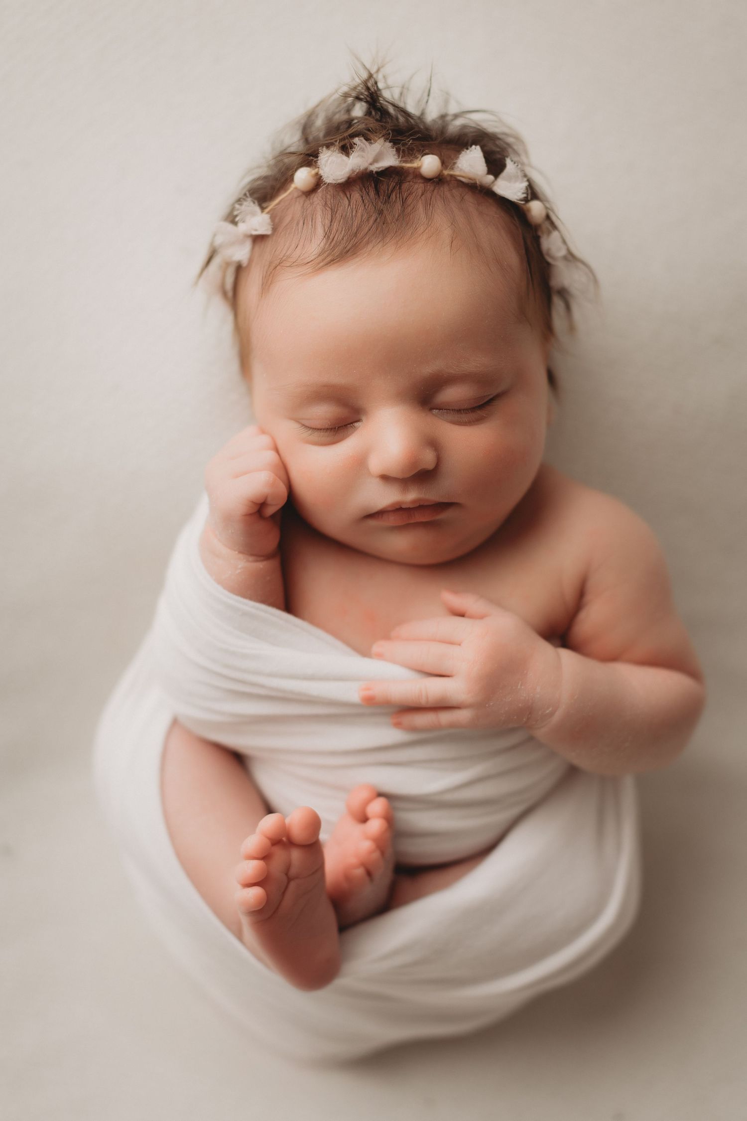 A peaceful newborn baby girl sleeps wrapped in white cloth wearing a delicate floral headband.