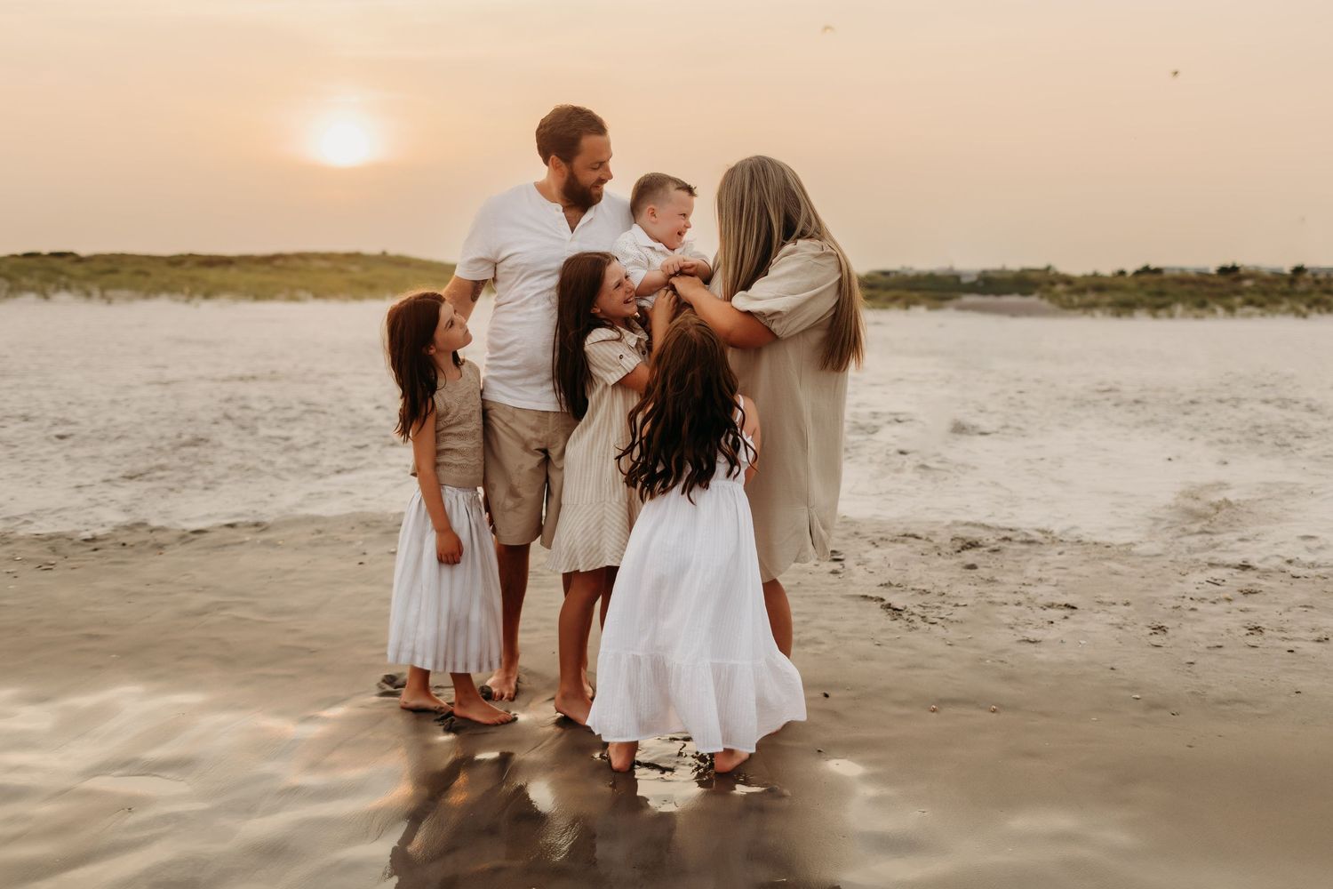 Family of five sharing a tender moment on a sandy beach at golden sunset, dressed in white and beige outfits.